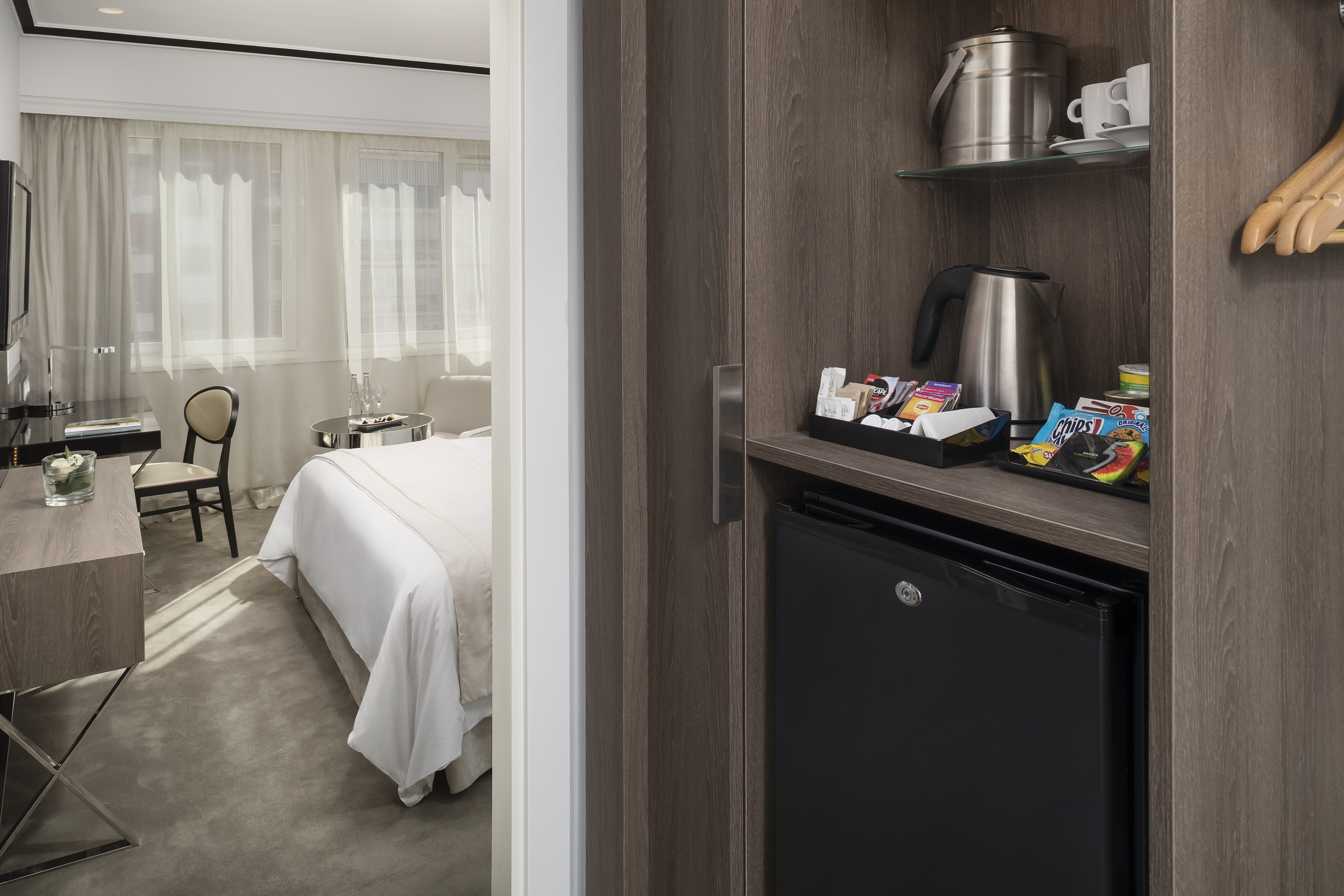 Hotel room minibar with fridge, kettle, coffee, snacks, and contemporary bedroom.