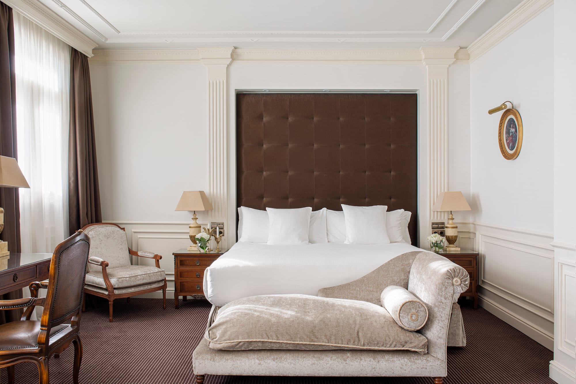 Elegant hotel bedroom with king bed, tufted headboard, and chaise lounge.