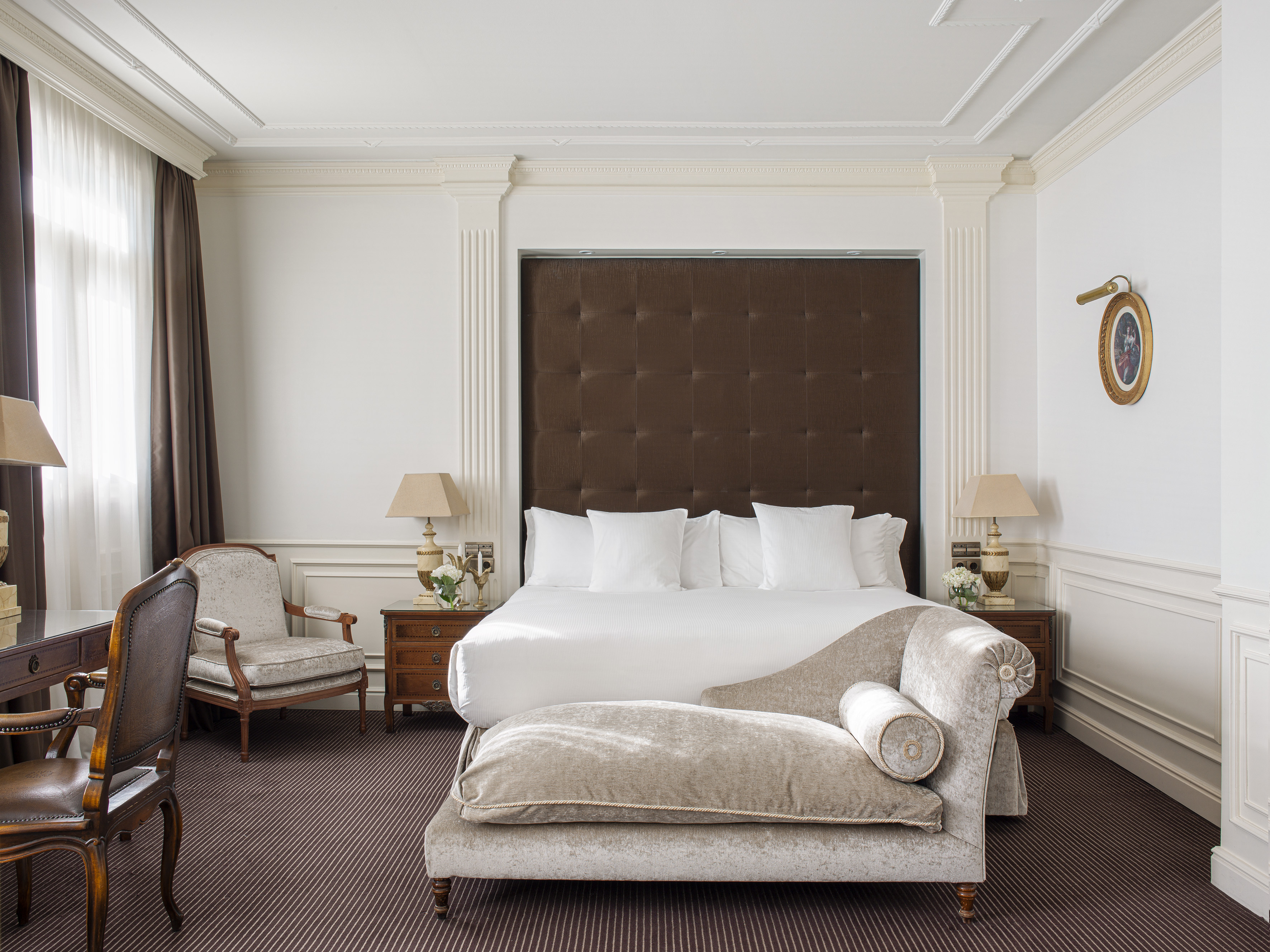Elegant hotel bedroom with king bed, tufted headboard, and chaise lounge.