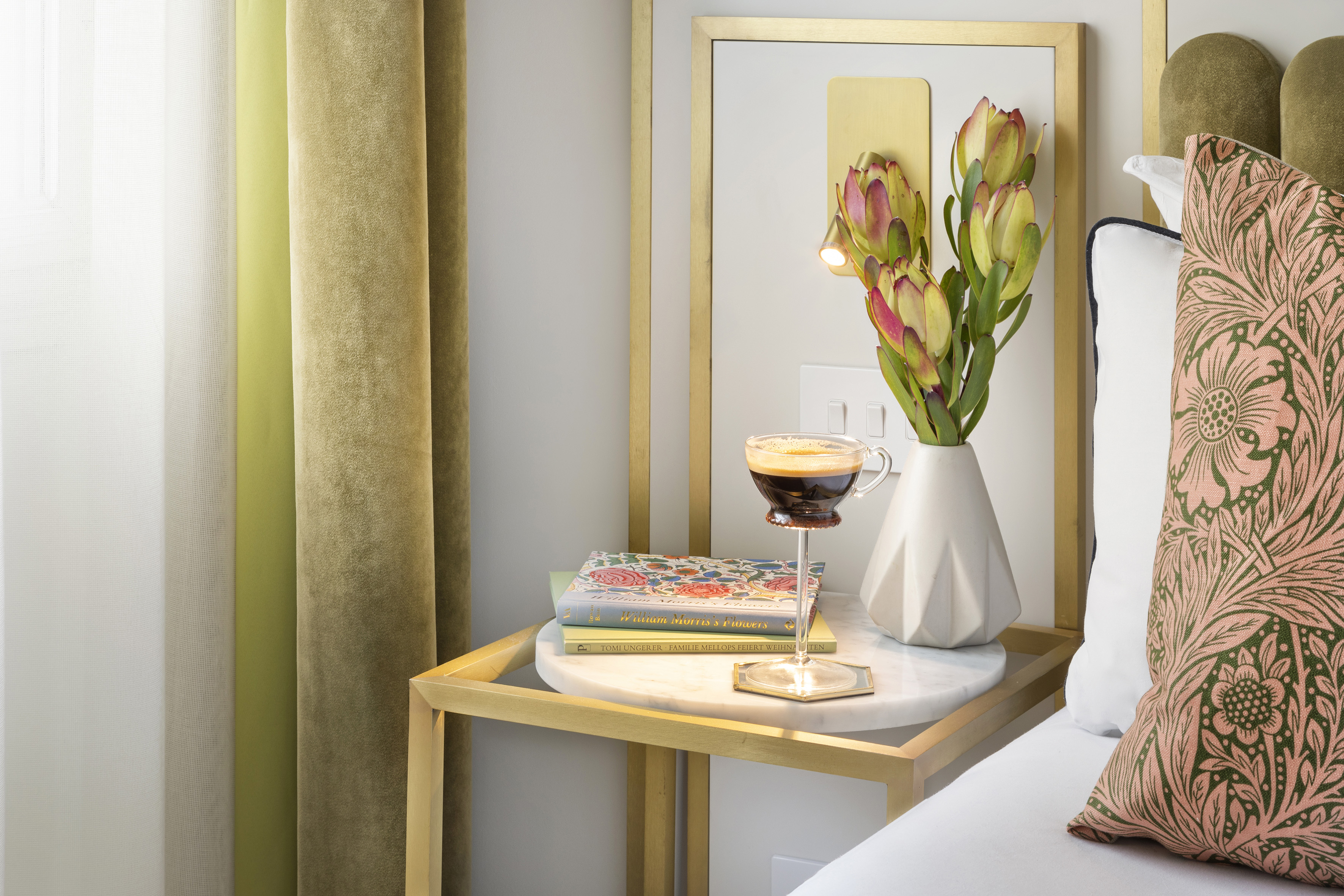 Elegant nightstand with coffee, floral books, protea vase, velvet curtains.