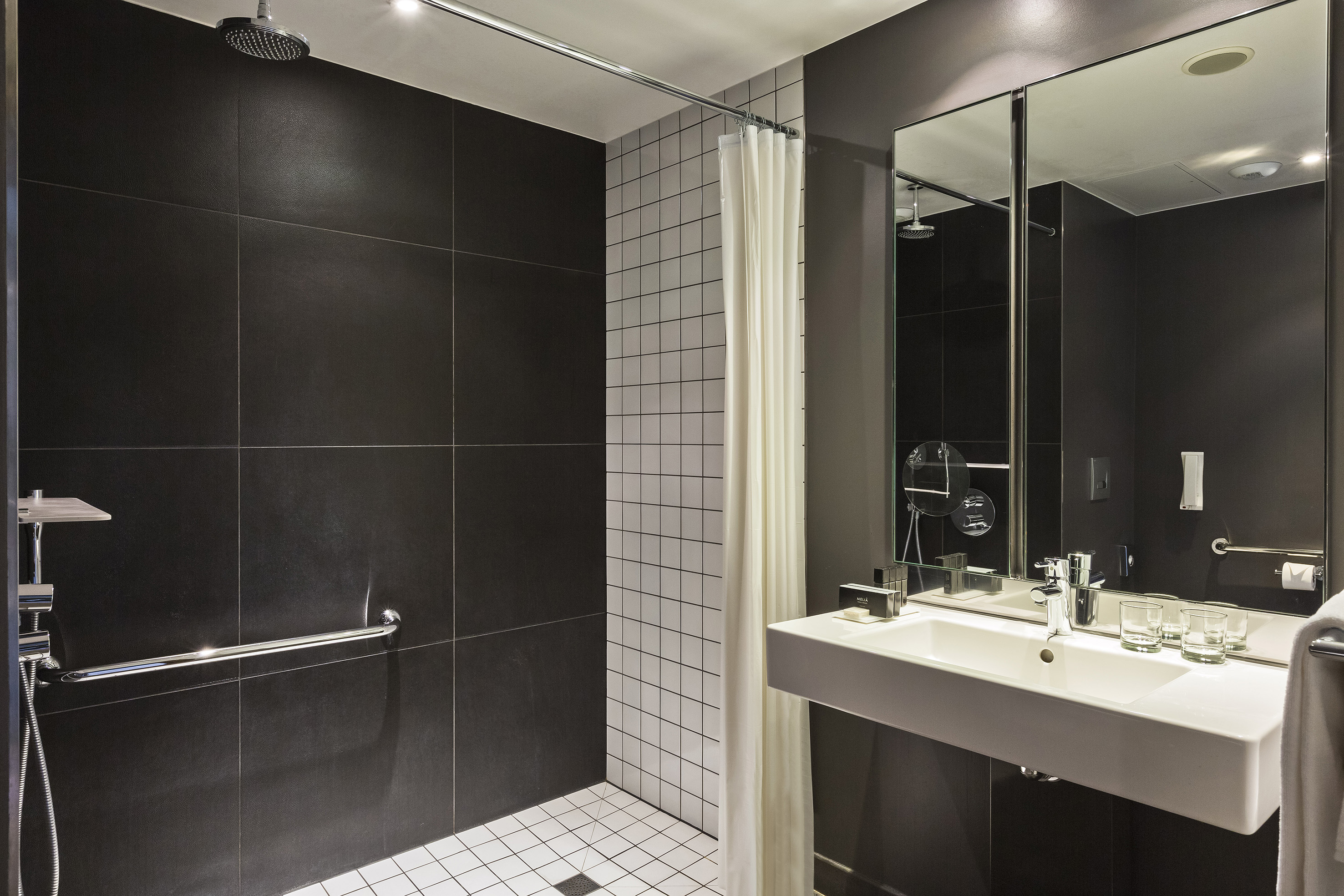 Modern hotel bathroom with dark tiled shower, white sink, and mirror.