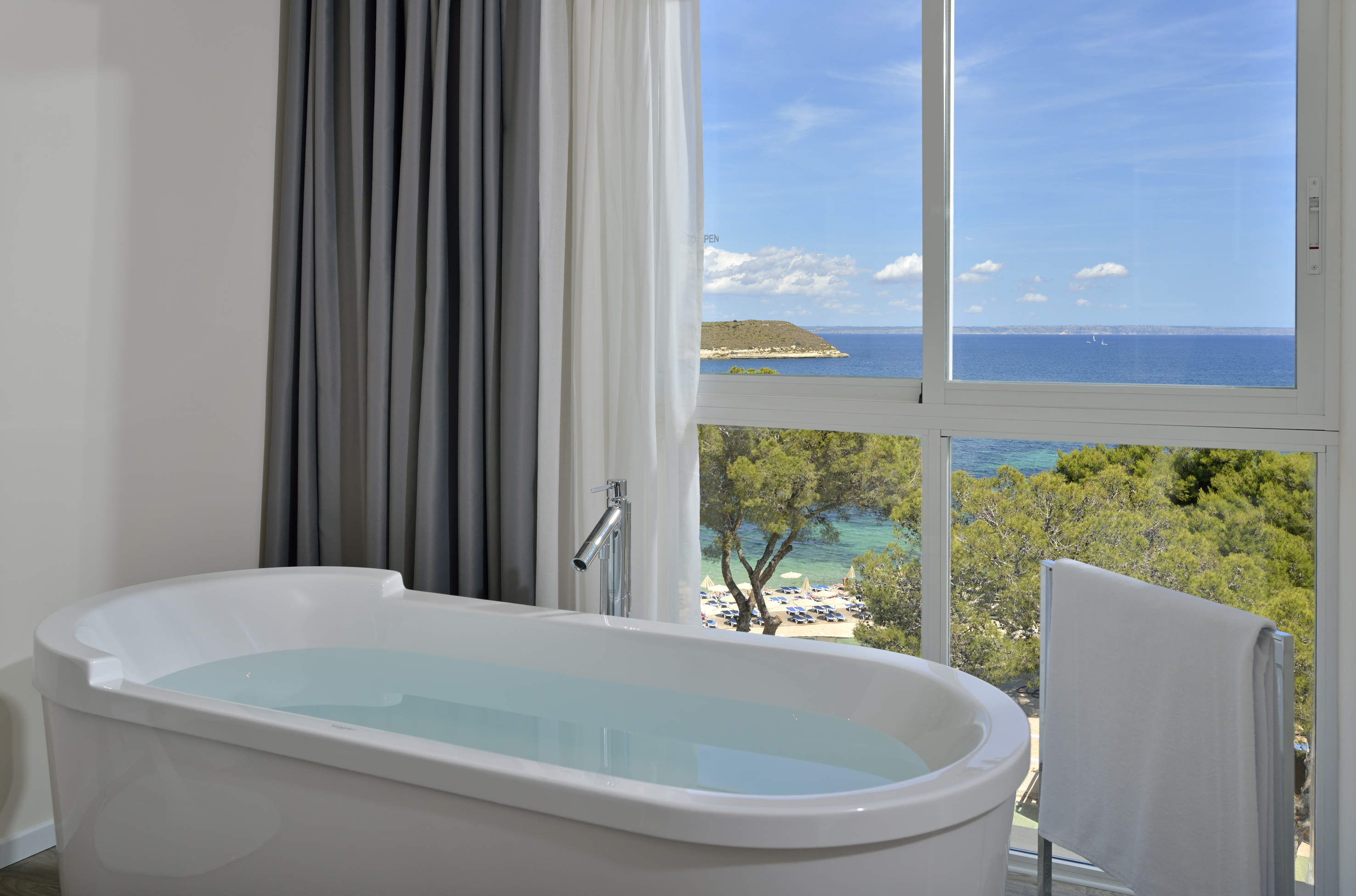 Bathtub with stunning ocean view, sandy beach below, and green trees.