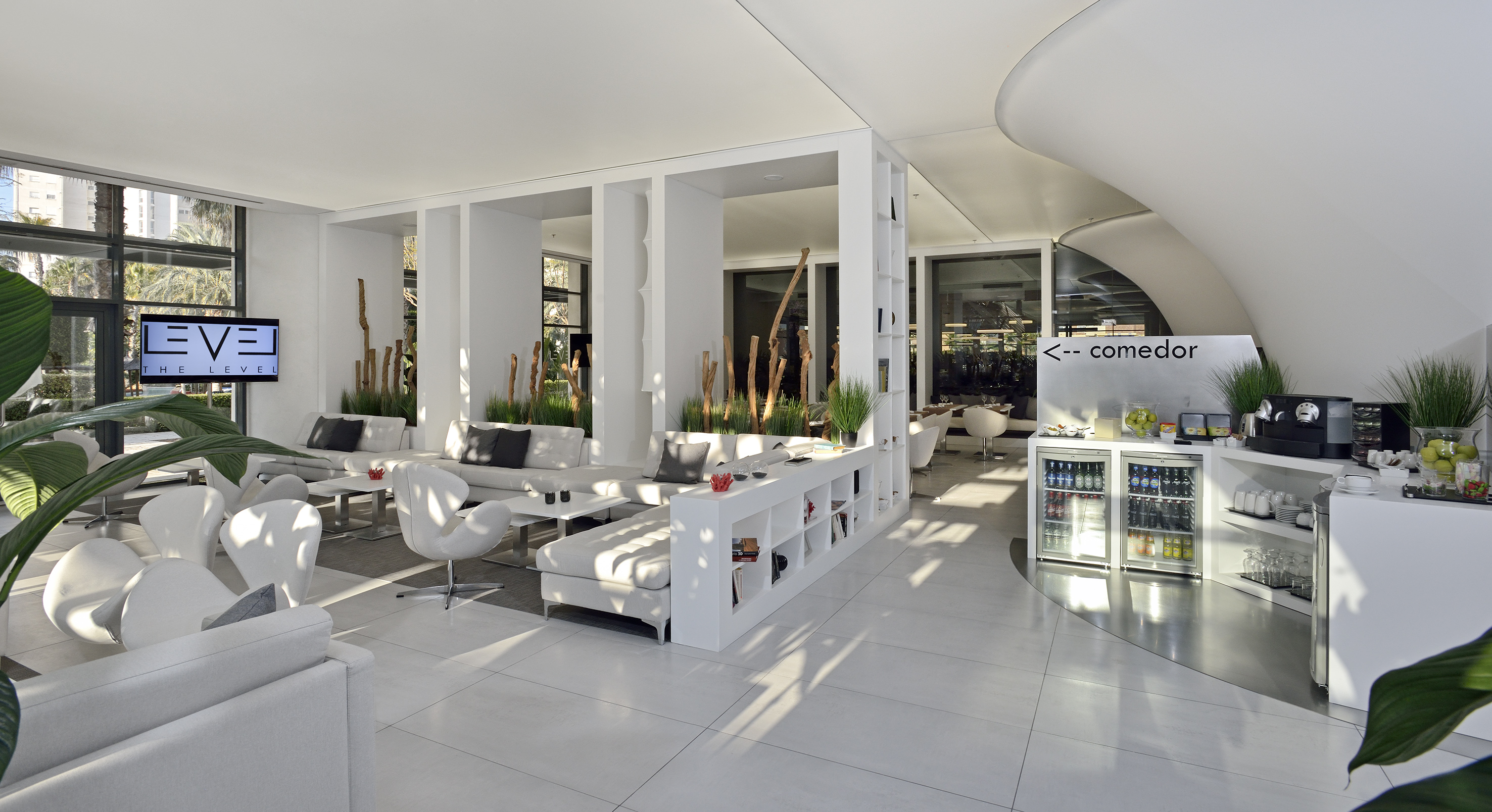 Bright modern hotel lounge with white seating, plants, and refreshment bar.