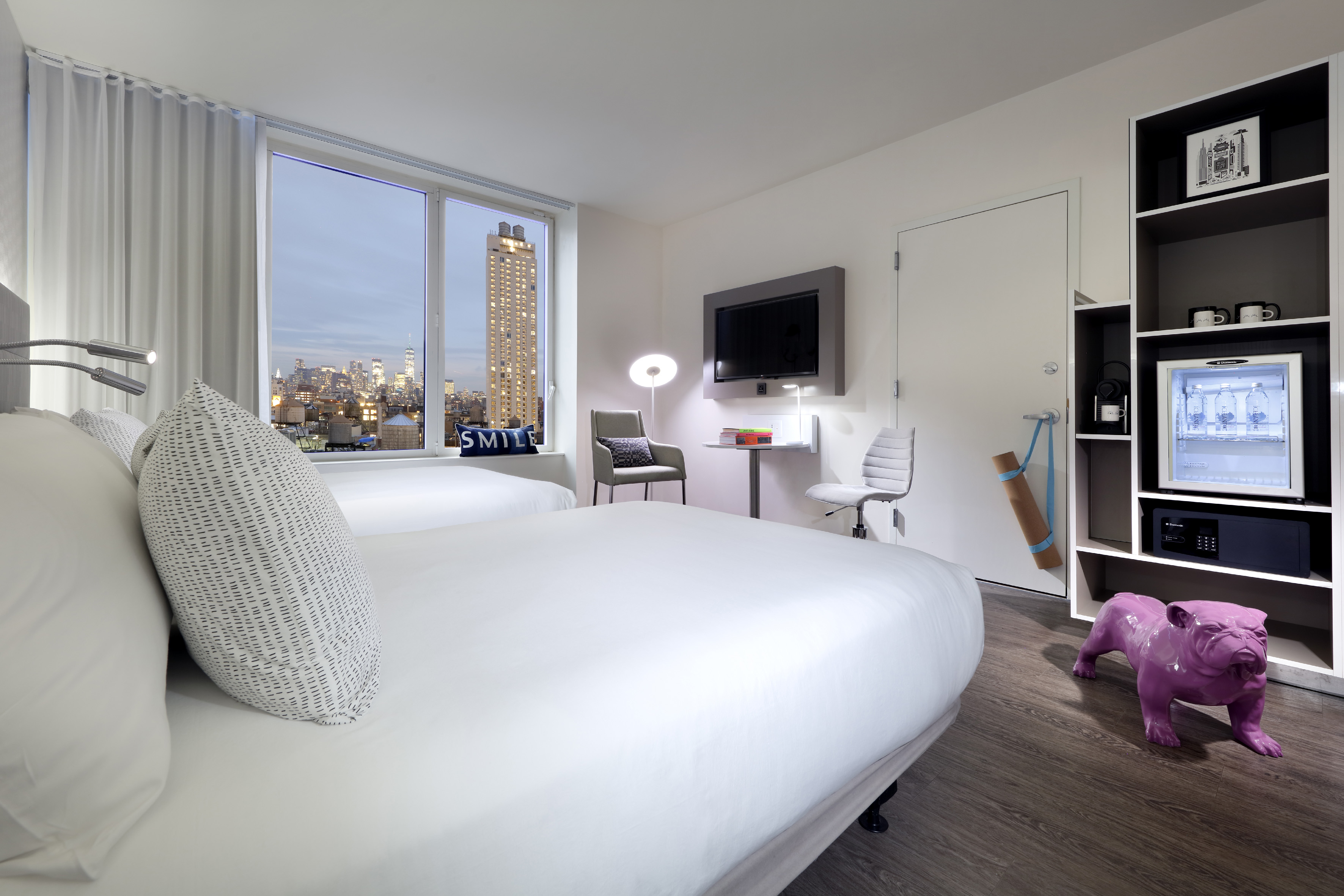 NYC hotel room with two beds, city view, minibar, and purple bulldog art.