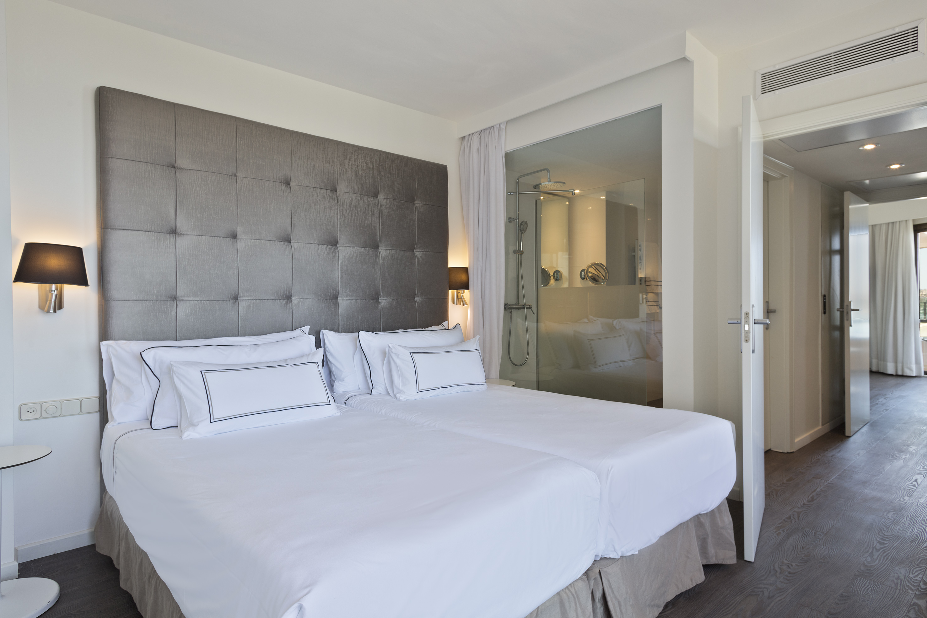 Modern hotel bedroom with twin beds, gray headboard, and glass shower partition.