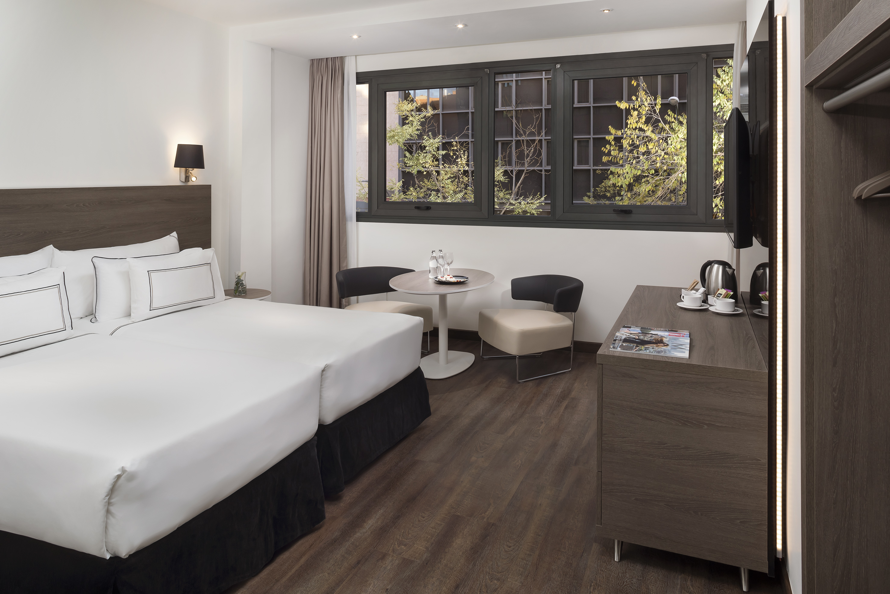 Modern hotel room with twin beds, dark wood flooring, and large window.