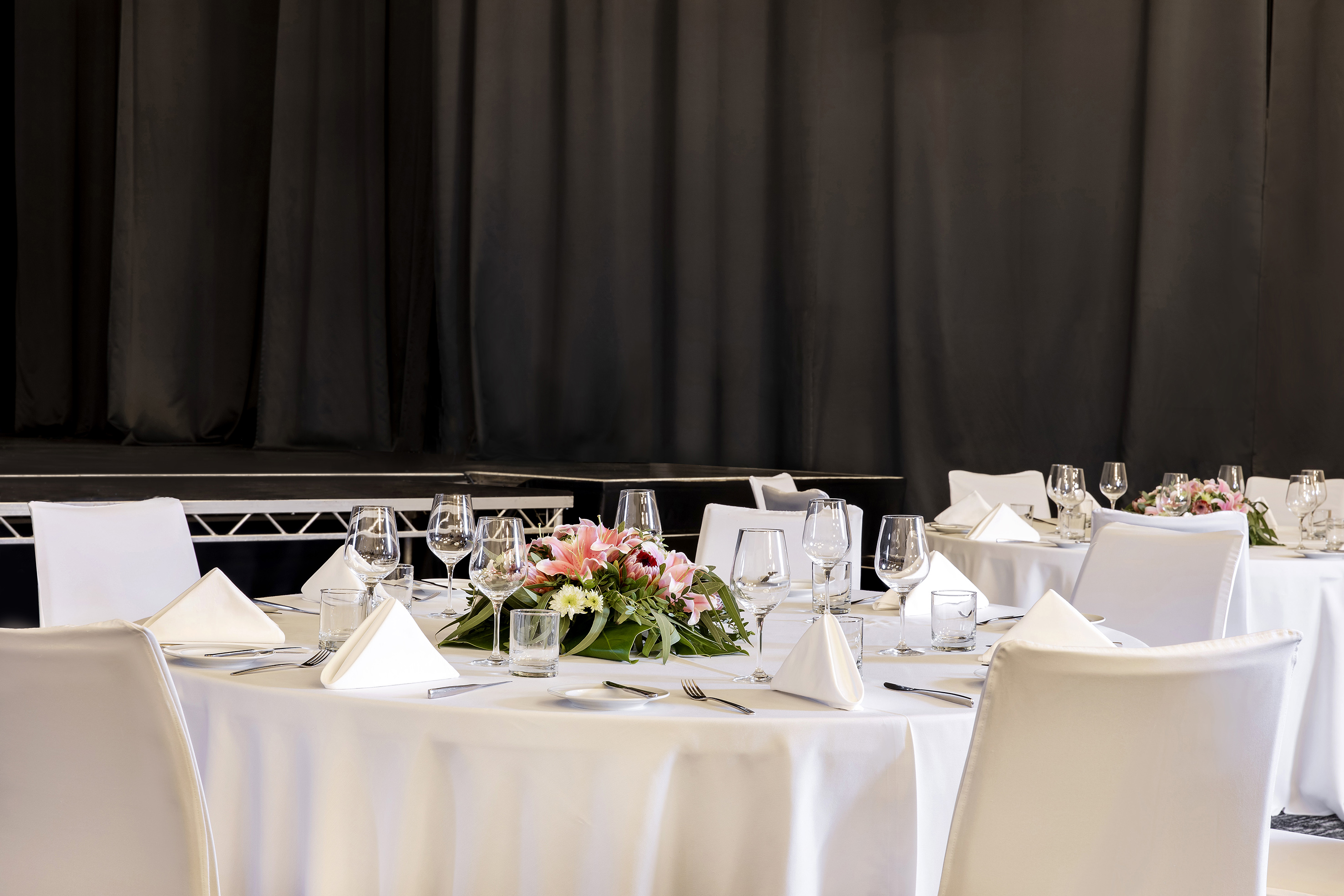 Conference dining tables with white settings, floral centerpieces, black stage.
