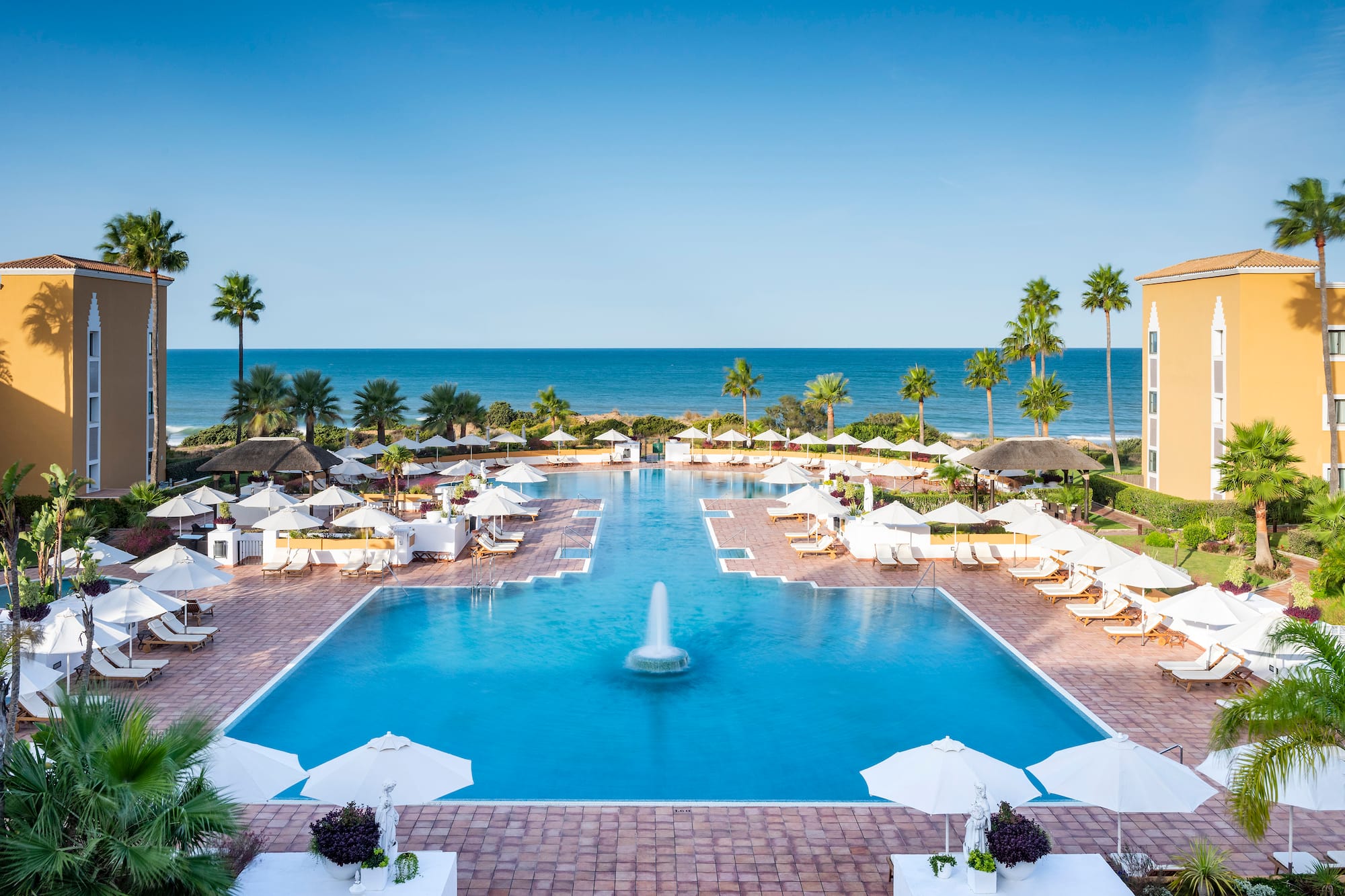 Luxury hotel resort pool with fountain, palm trees, ocean view, and sun loungers.