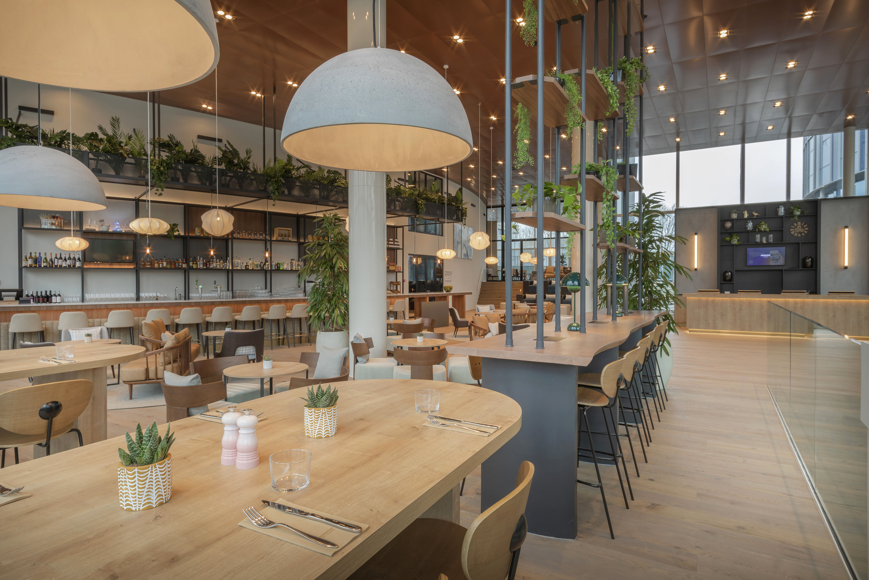 Modern hotel restaurant interior with concrete pendant lights, wooden tables, and green plants.