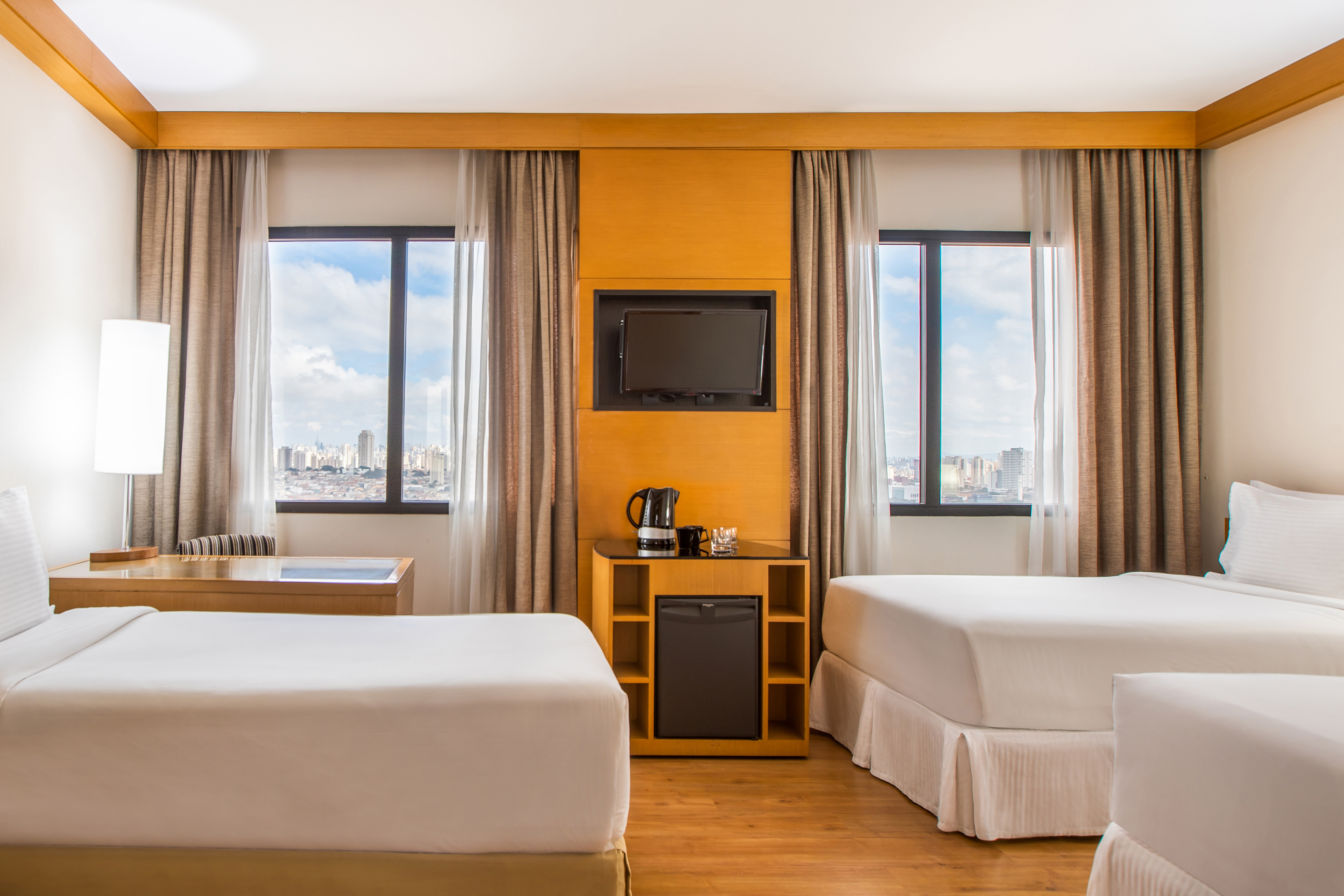 Bright hotel room with twin beds, city skyline view, TV, and mini-fridge.