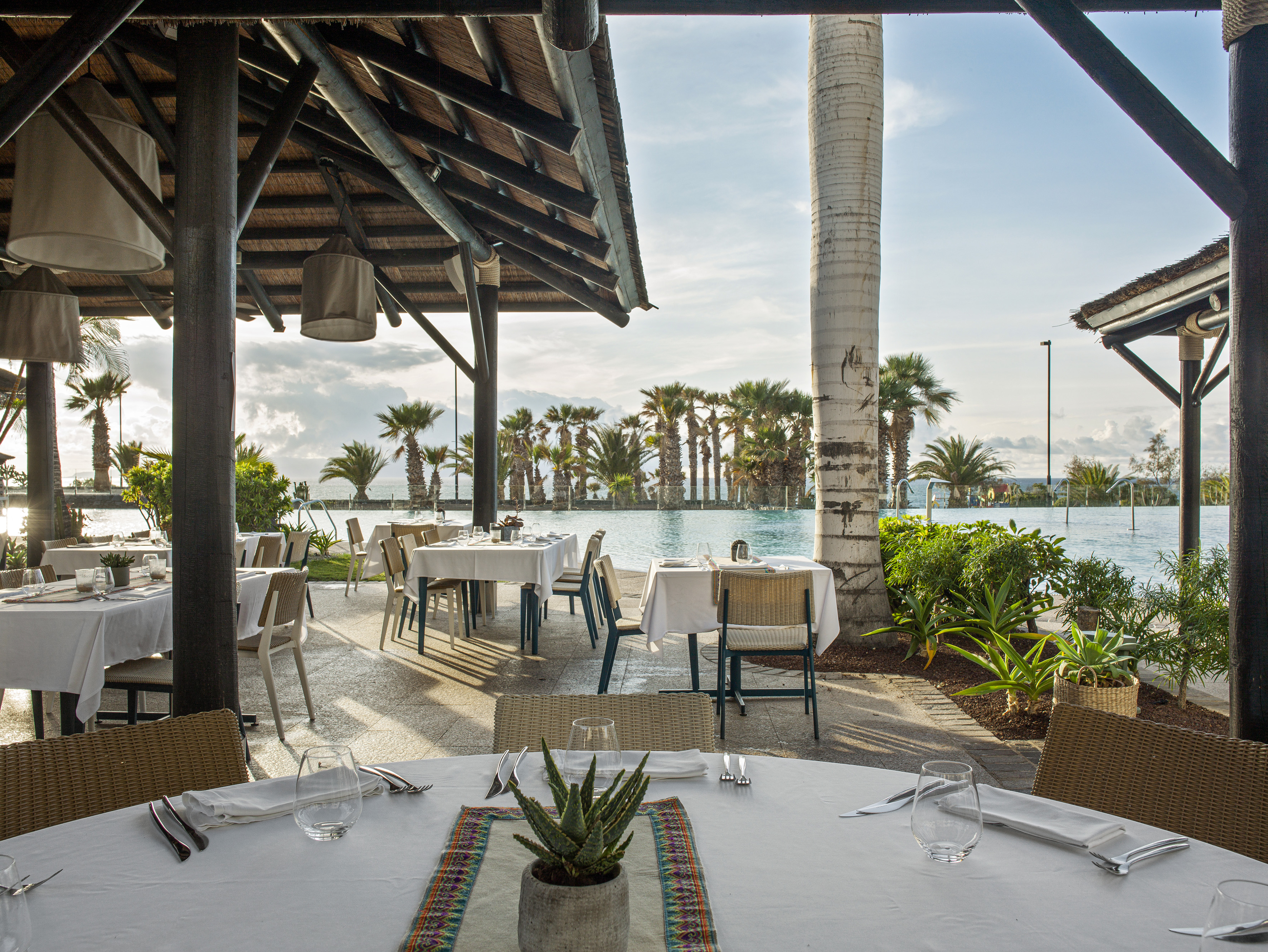 Al fresco dining at tropical resort, thatched roof, tables, infinity pool, palm trees.