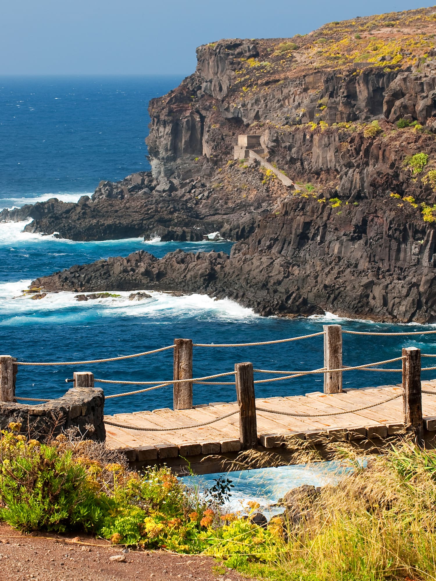 Coastal boardwalk with rope railing, rugged cliffs, and blue ocean waves.