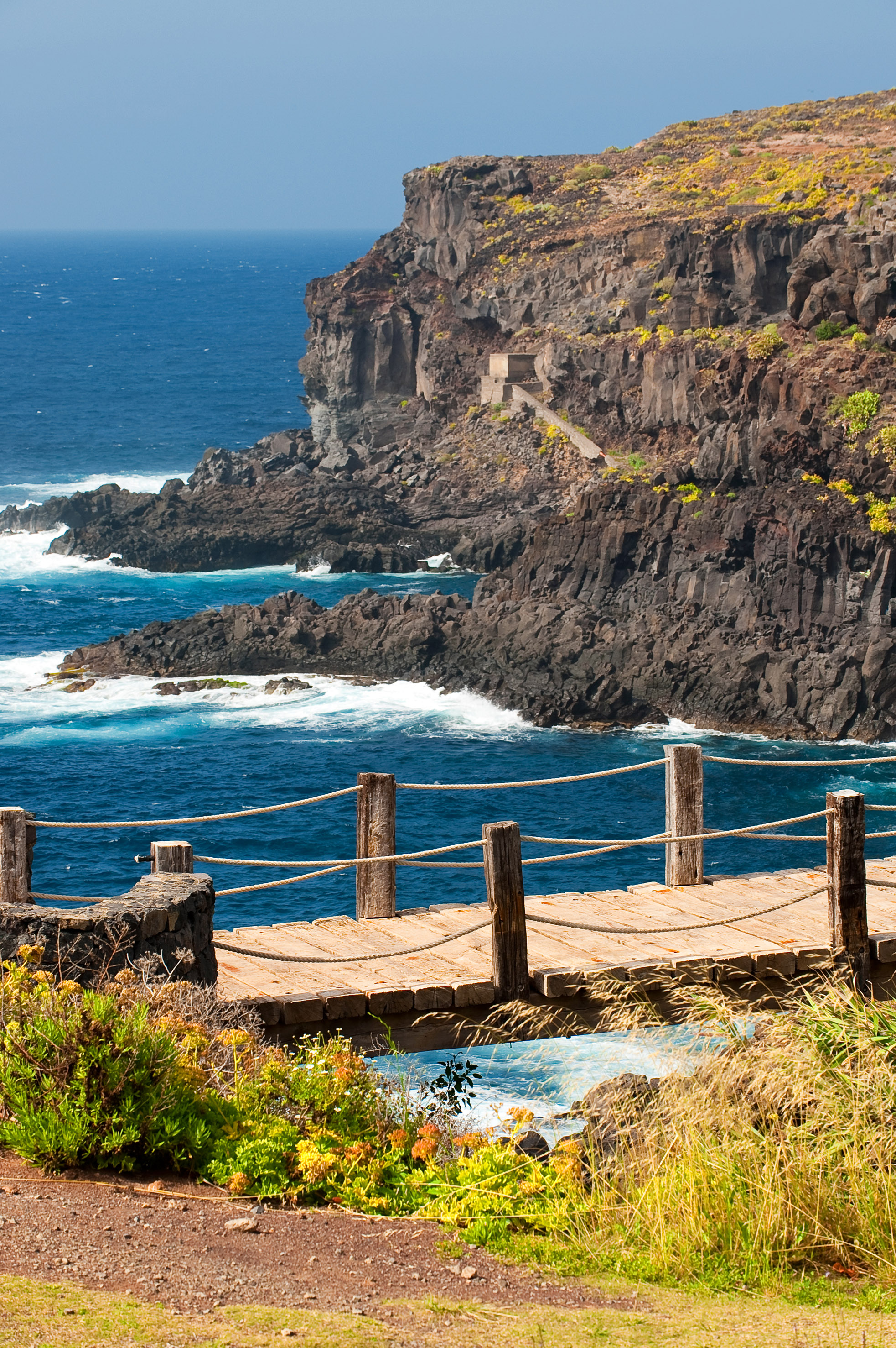 Coastal boardwalk with rope railing, rugged cliffs, and blue ocean waves.