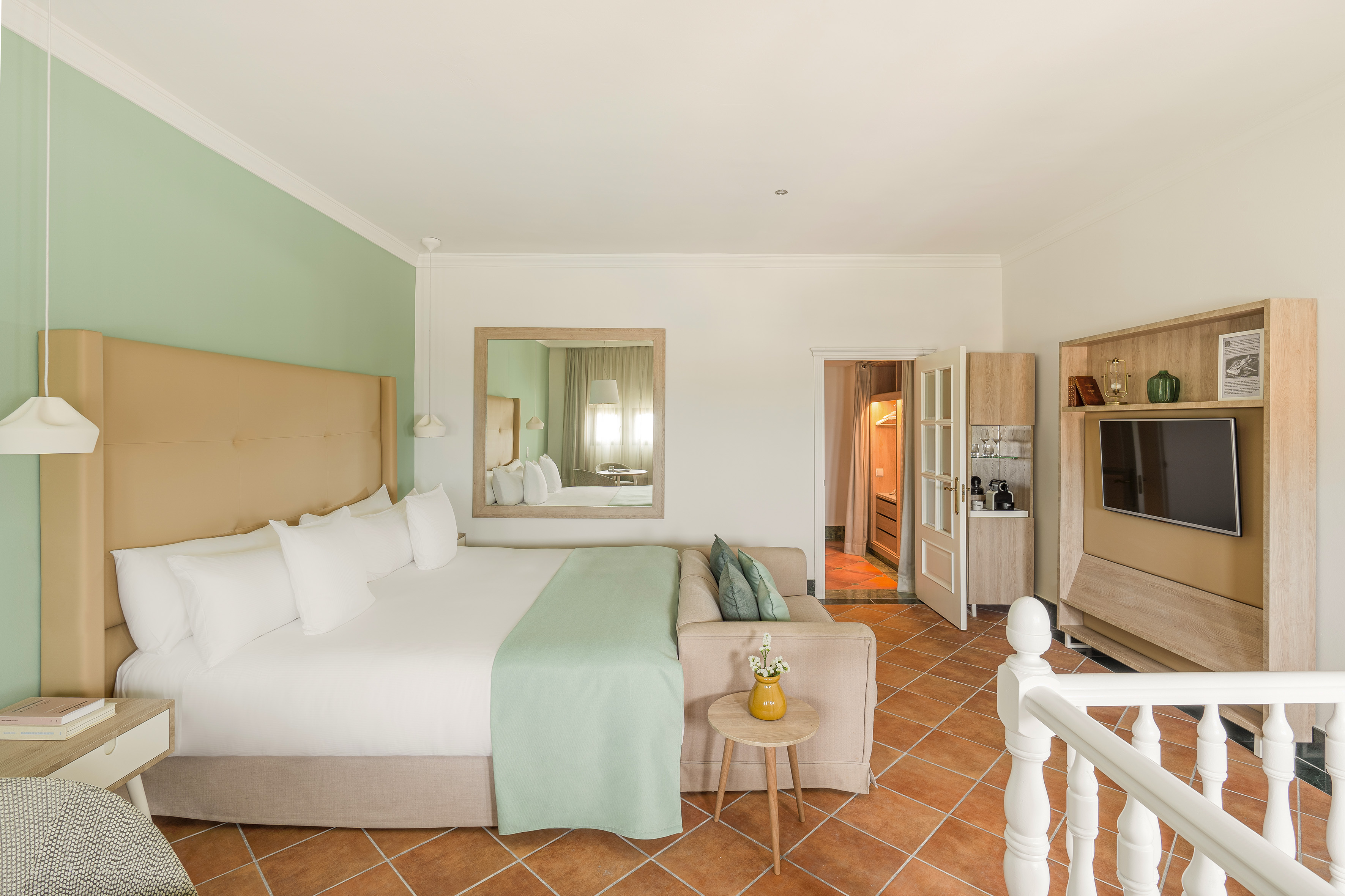 Modern hotel room with king bed, green accents, sofa, TV, and terracotta tiles.