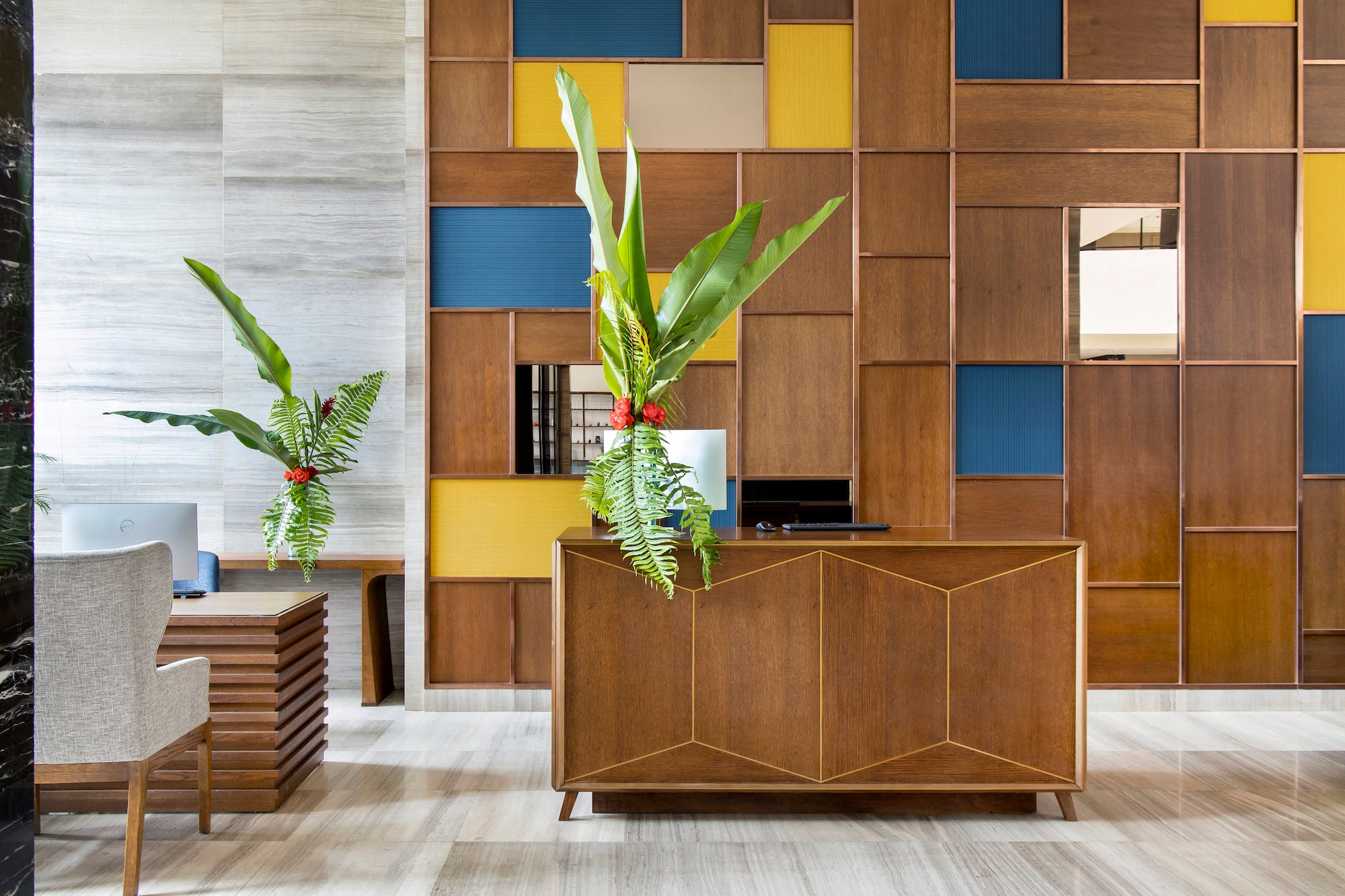 Modern hotel reception desk, wood panel wall with blue/yellow, tropical plants.