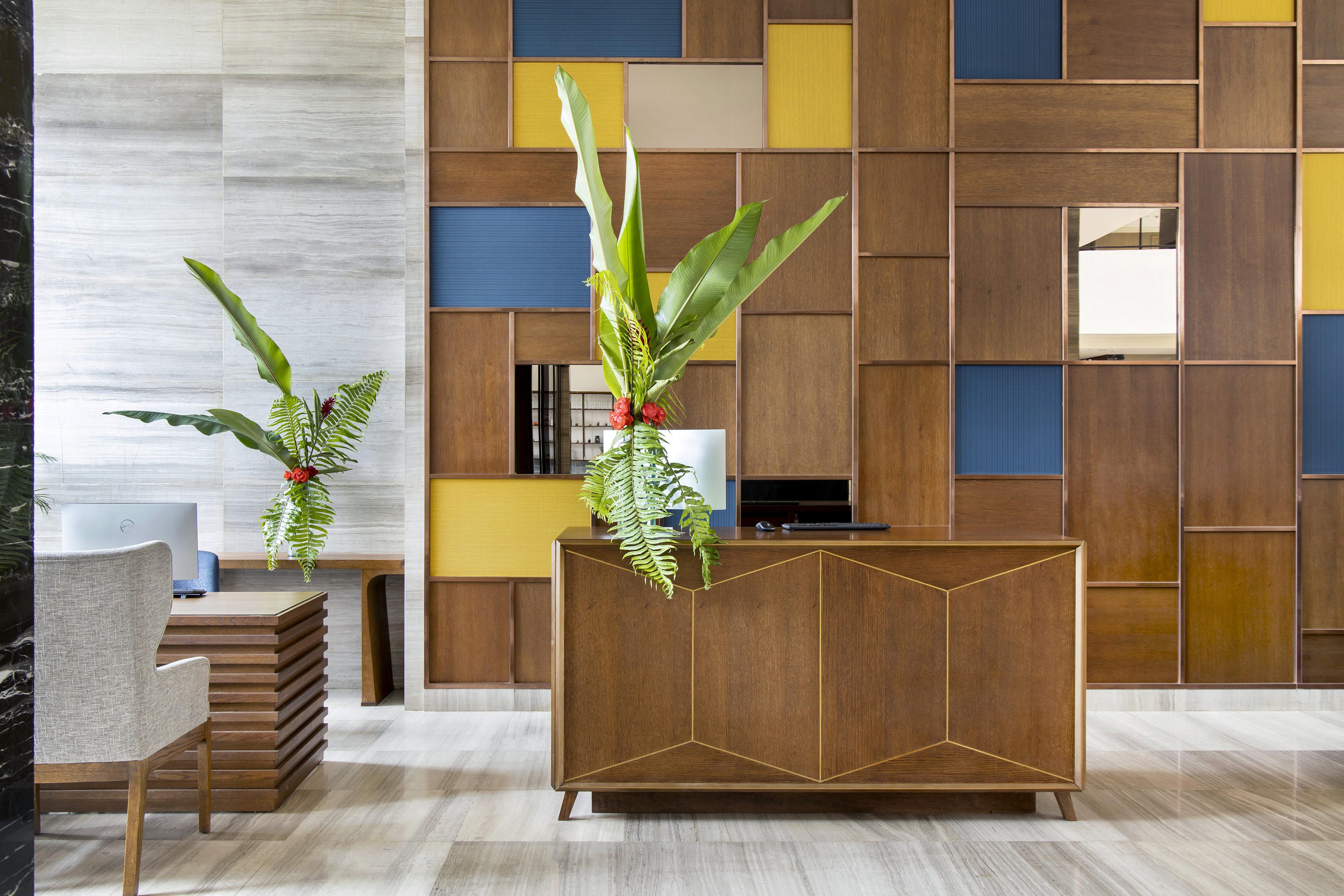 Modern hotel reception desk, wood panel wall with blue/yellow, tropical plants.