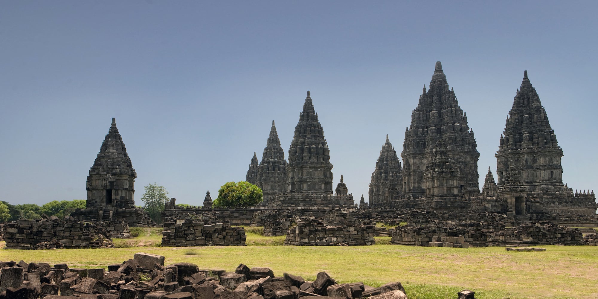 a stone structure with towers and grass with Prambanan in the background