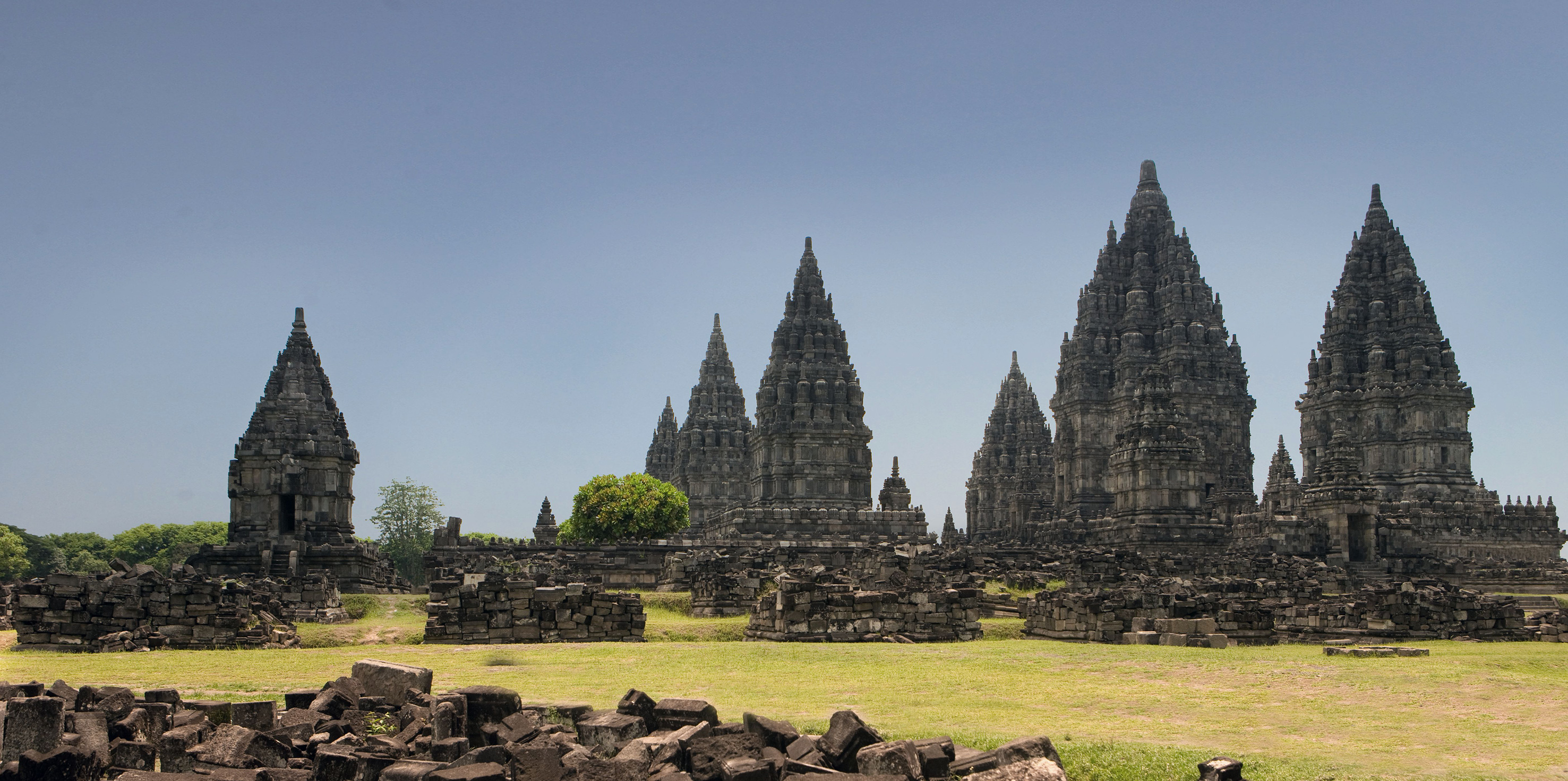 a stone structure with towers and grass with Prambanan in the background
