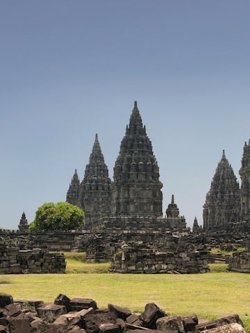 a stone structure with towers and grass with Prambanan in the background