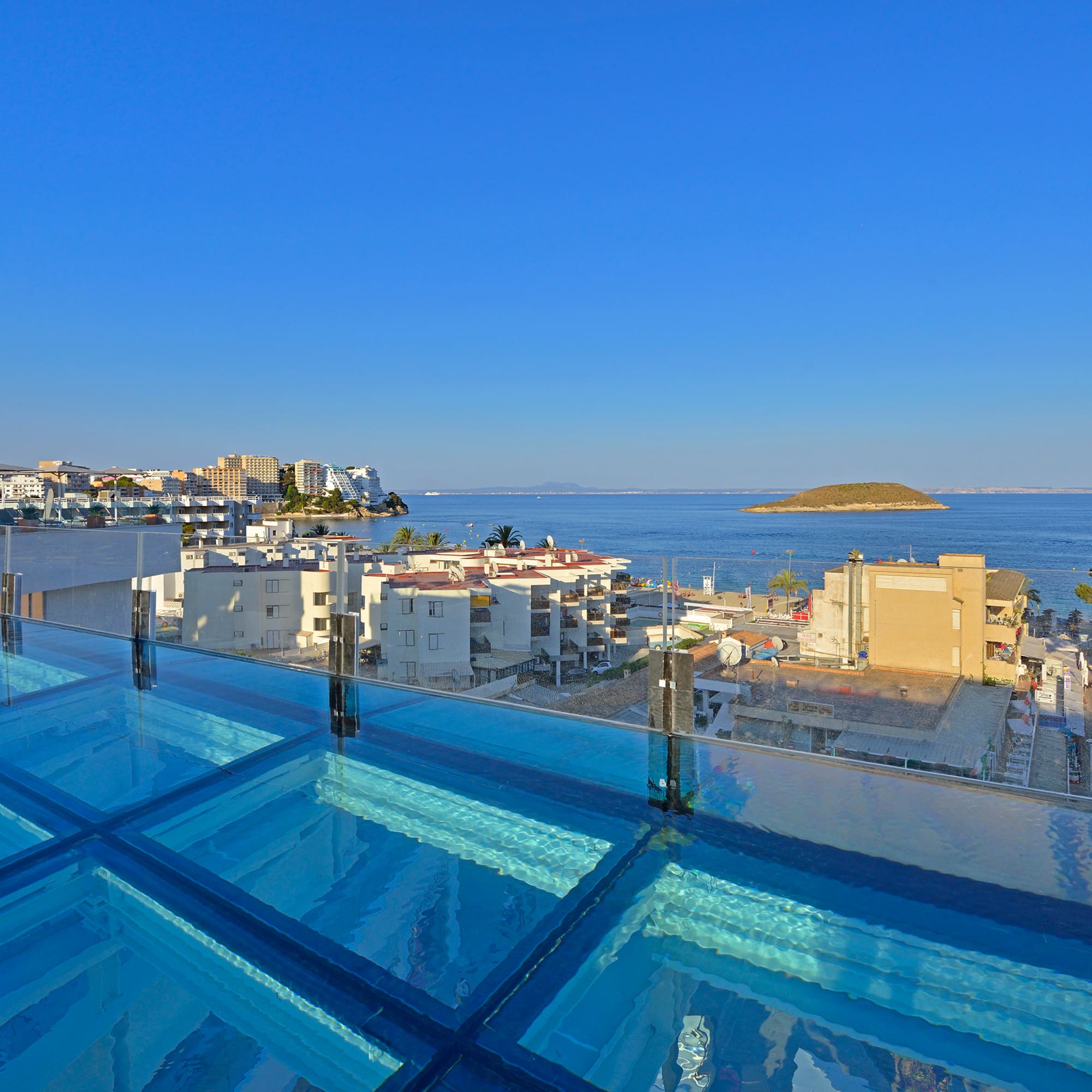 Rooftop glass pool overlooking a Mediterranean coastal town and distant island.