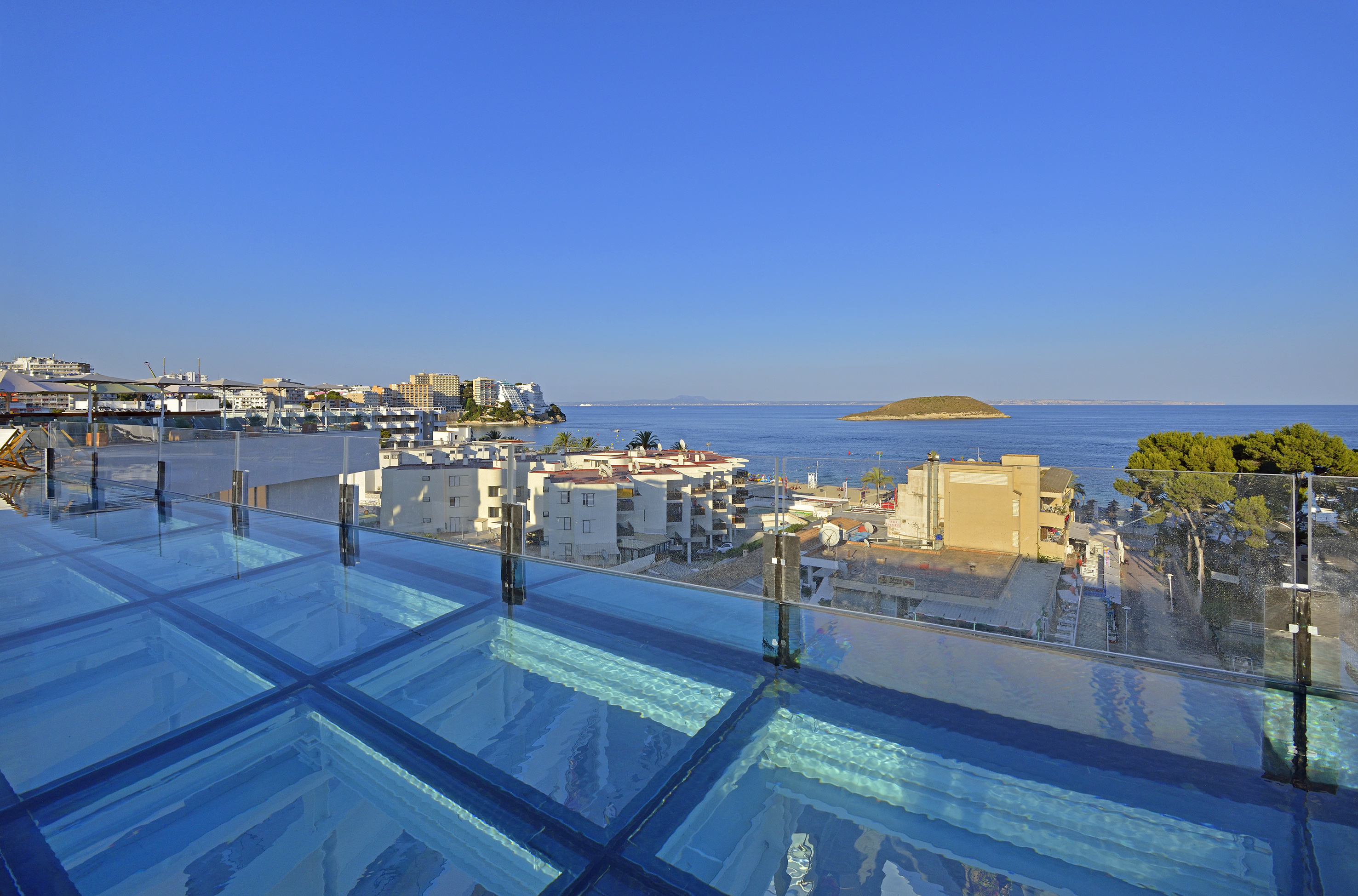 Rooftop glass pool overlooking a Mediterranean coastal town and distant island.