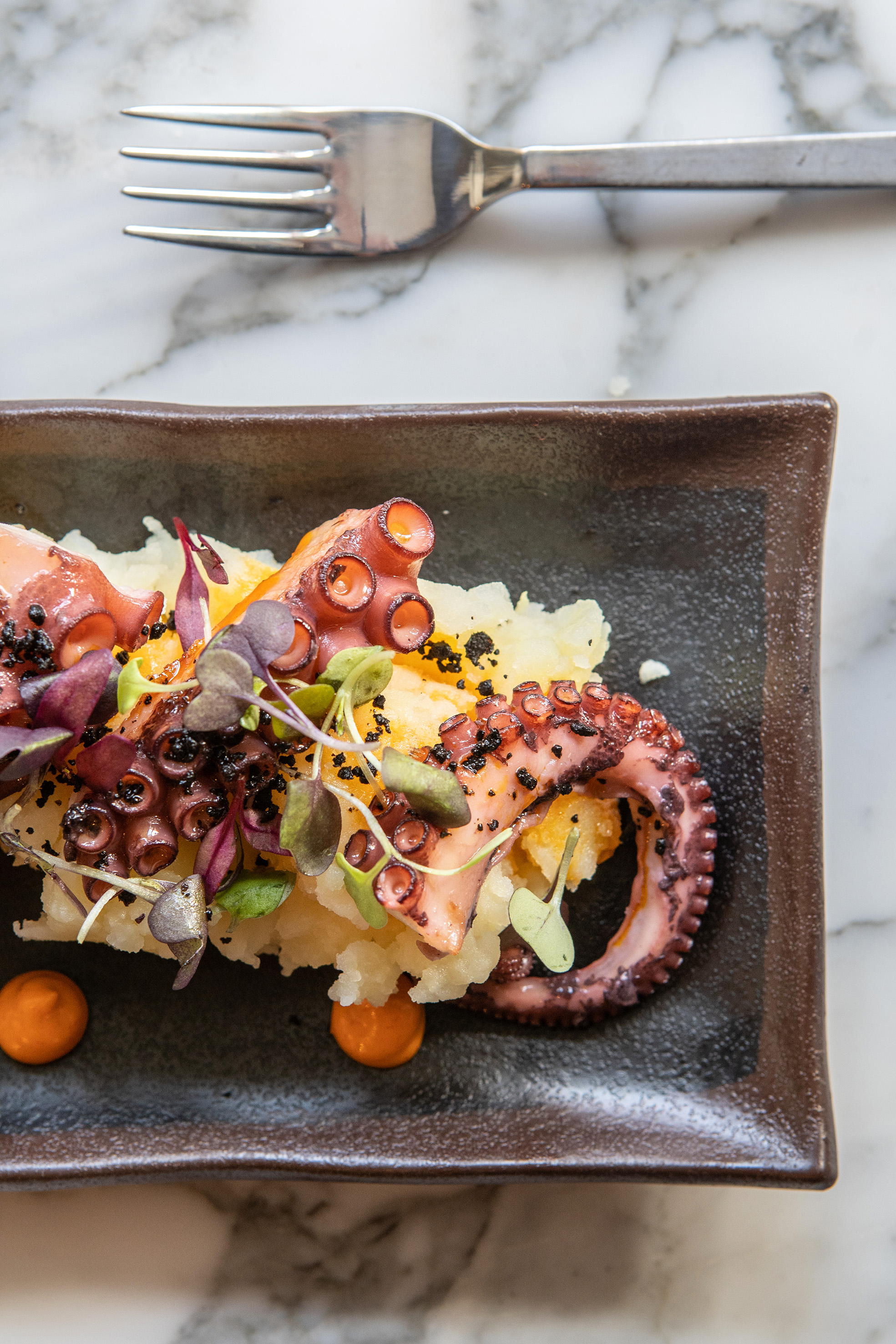 Octopus with mashed potatoes, microgreens, and orange sauce on a dark plate.