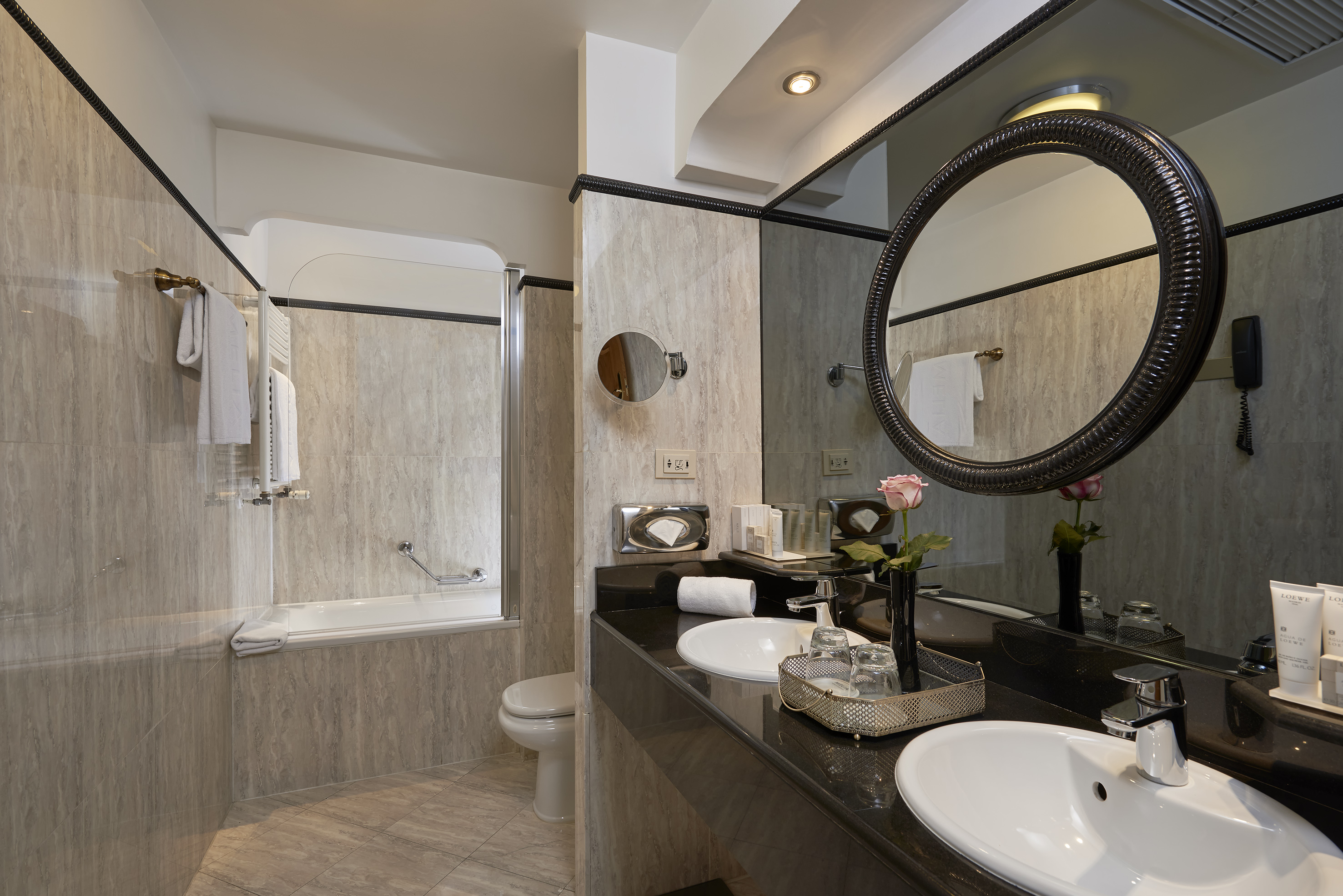 Luxury hotel bathroom with double vanity, ornate mirror, bathtub, and tiled walls.