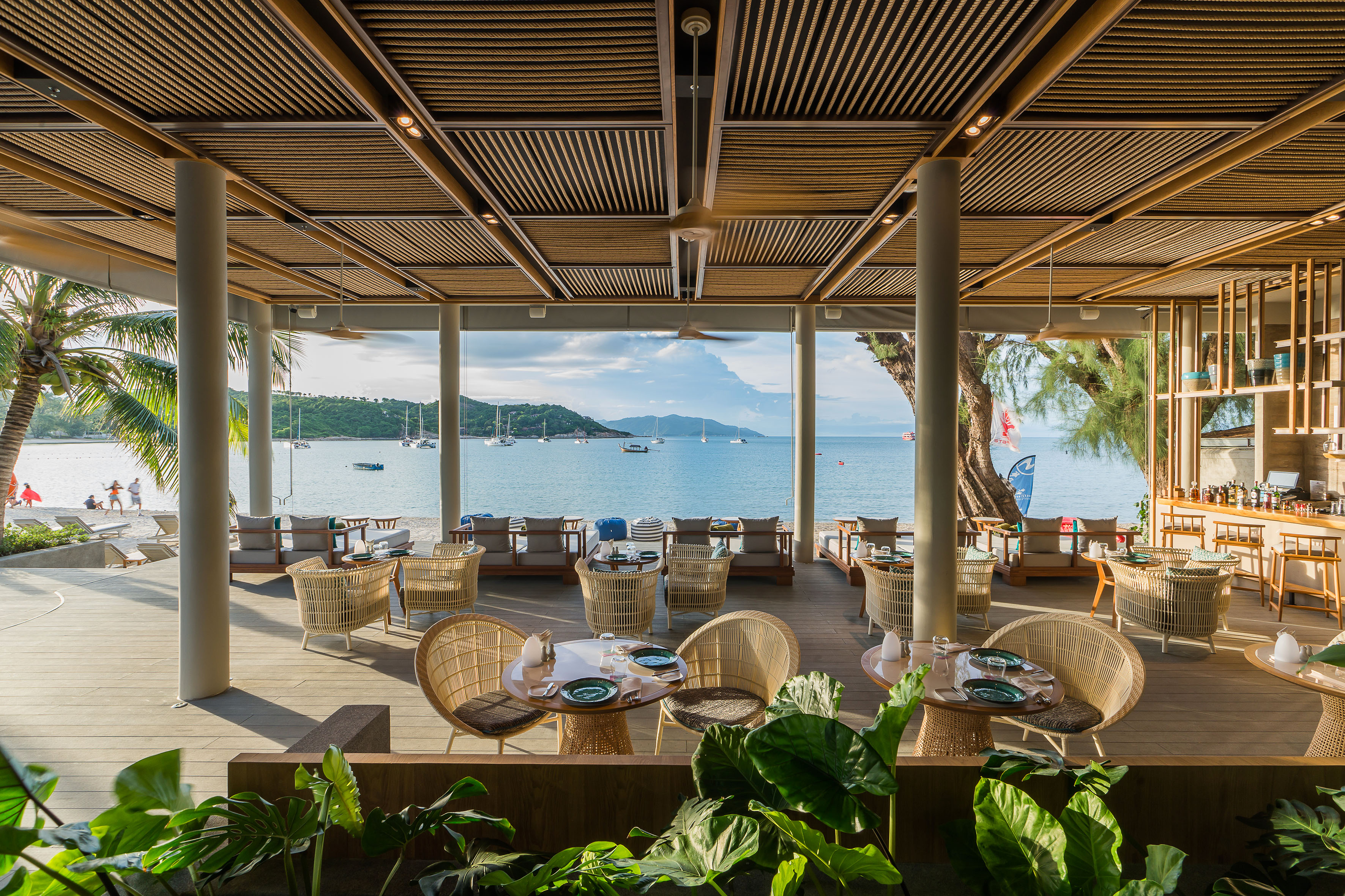 Beachfront restaurant with ocean view, wicker chairs, and boats in the bay.