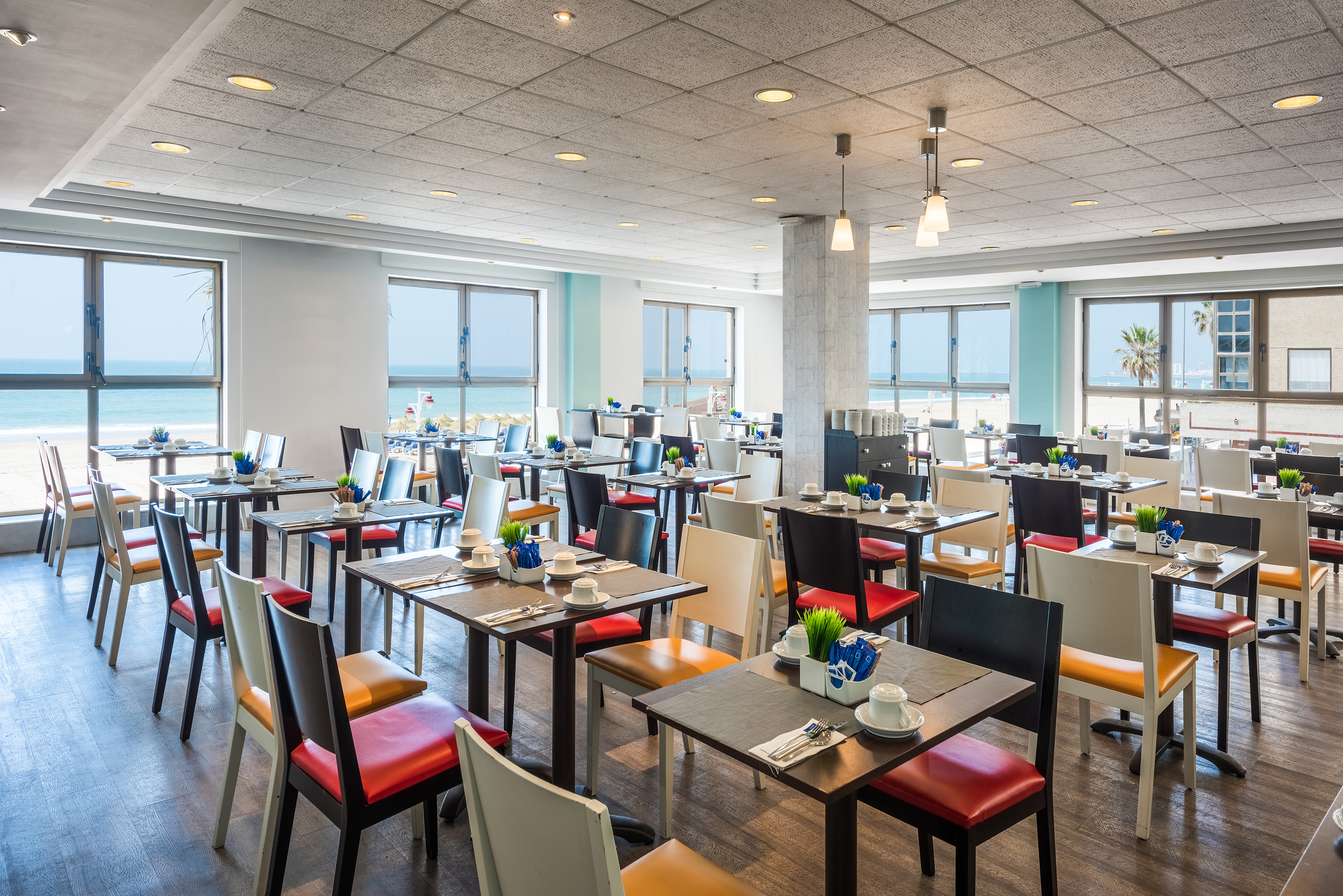 Modern restaurant with ocean view, set tables, and colorful dining chairs.