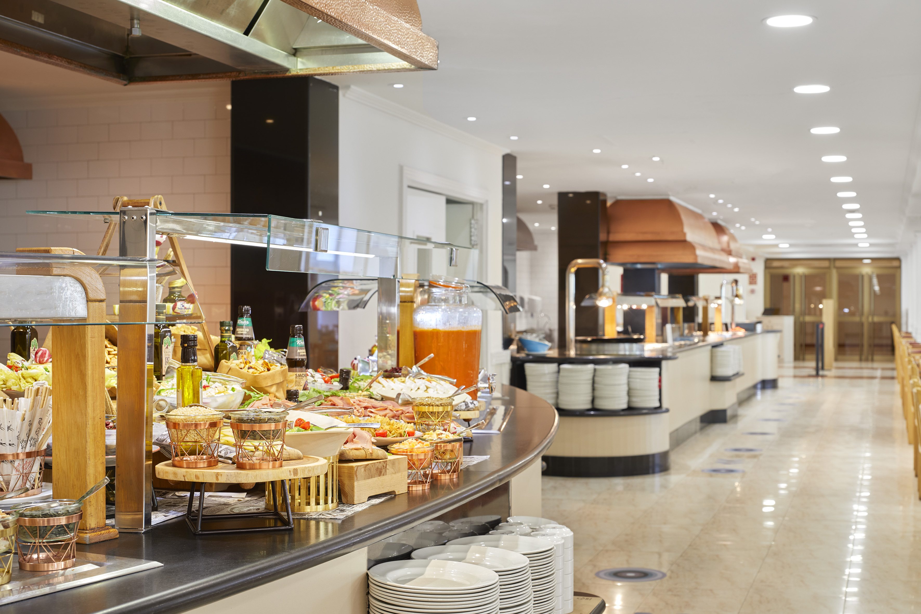 Spacious hotel buffet line with salads, cold cuts, juice, and stacked plates.