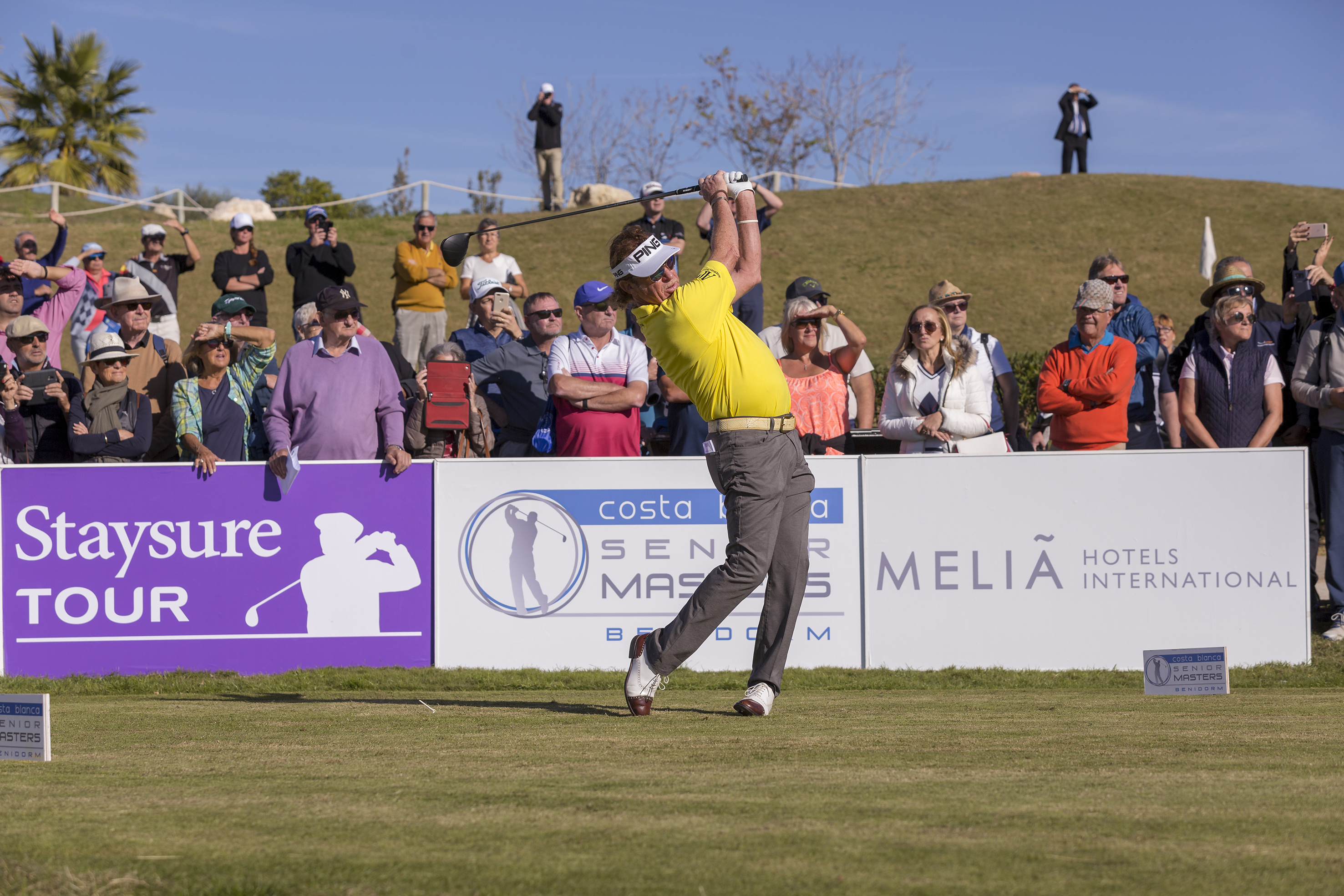 Golfer in yellow shirt swings driver at Staysure Tour Senior Masters with crowd watching.