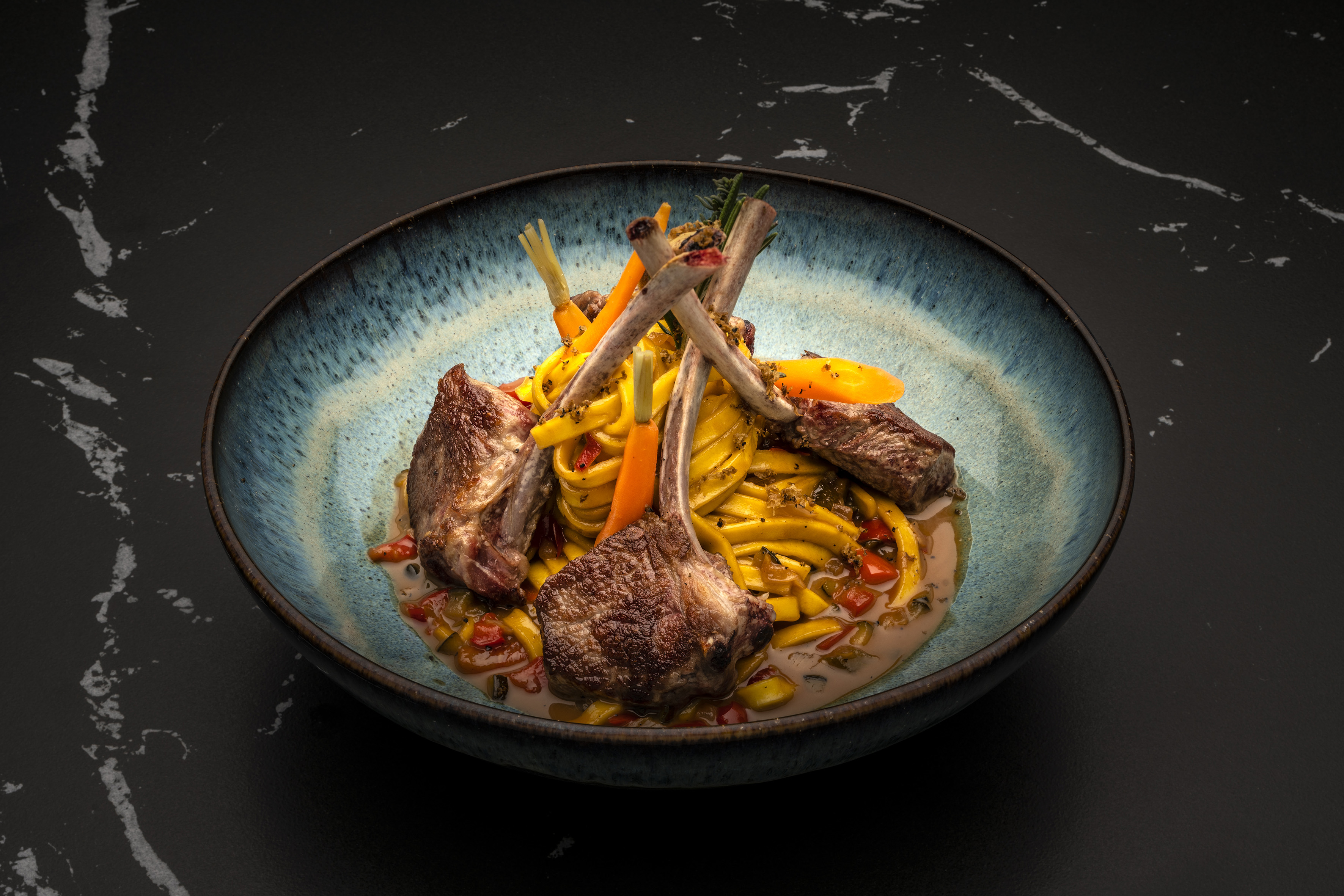 Seared lamb chops with fresh pasta, baby carrots, and rich sauce in blue bowl.