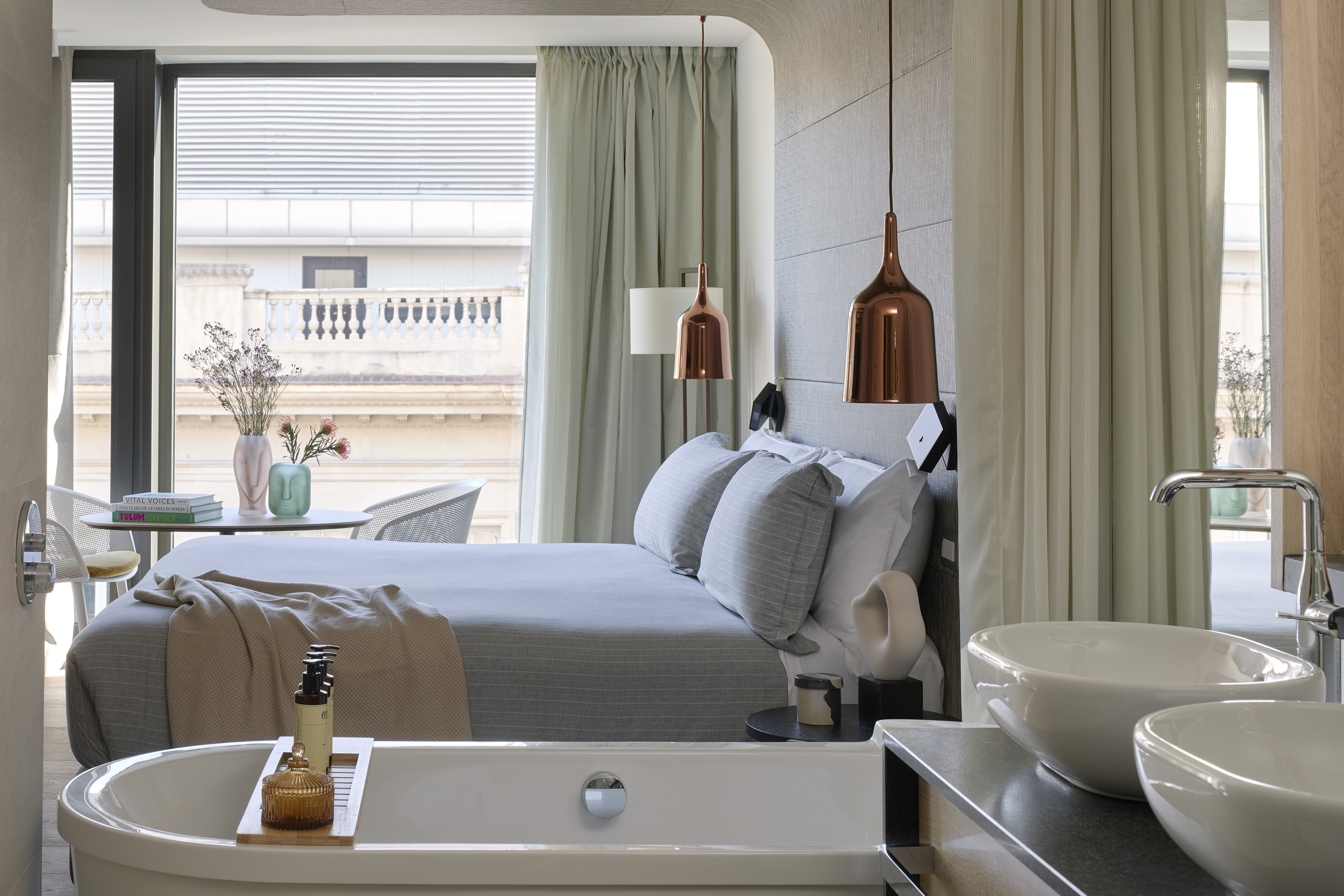 Modern hotel suite with bed, freestanding tub, dual sinks, and city view.