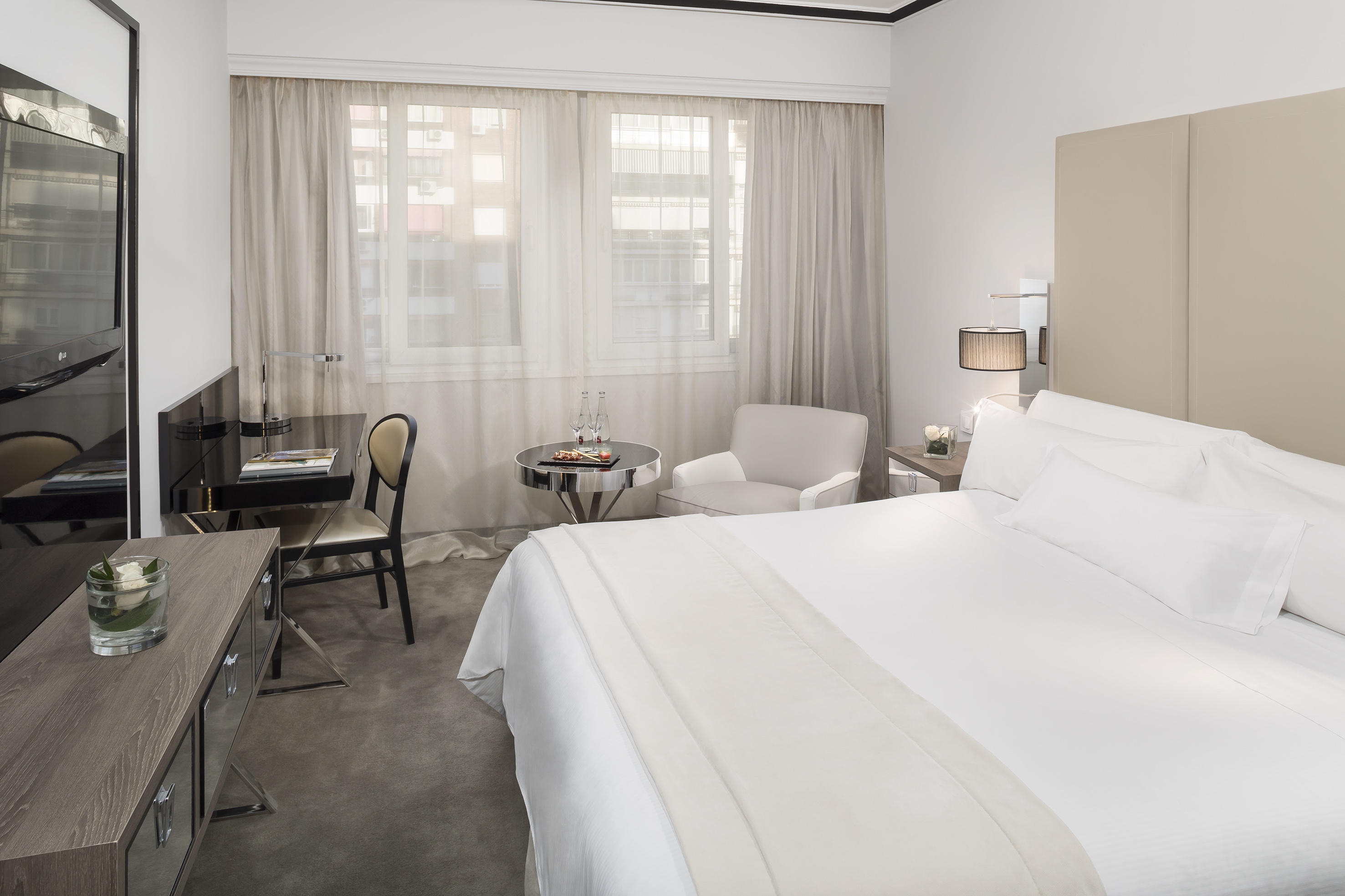 Modern hotel bedroom with white bed, work desk, armchair, and window.