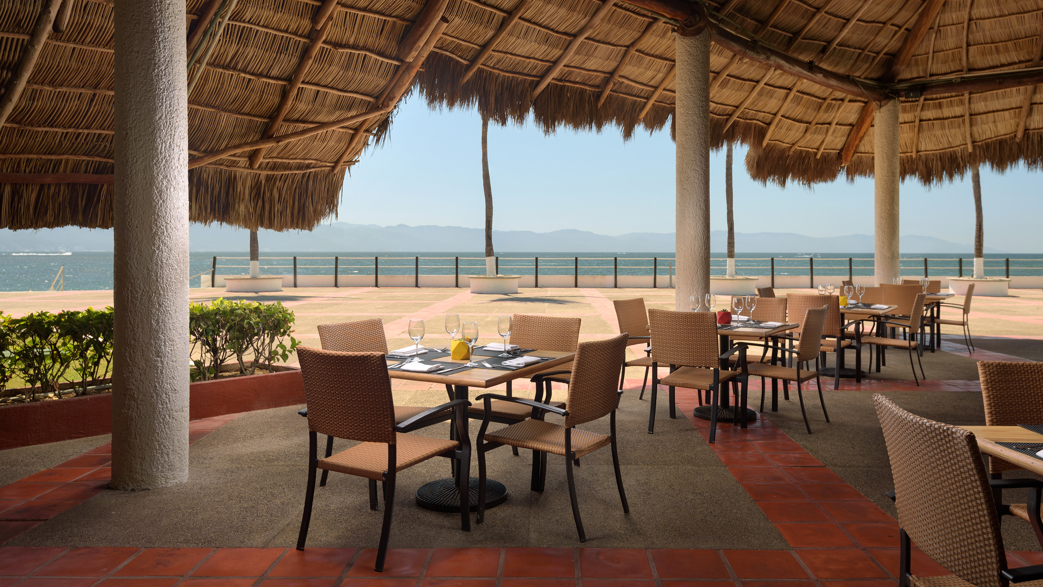 Outdoor ocean view restaurant with palapa roof and set tables.