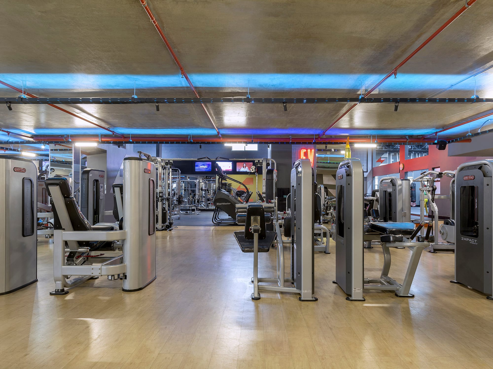 Gym interior with diverse strength and cardio workout machines under blue lighting.