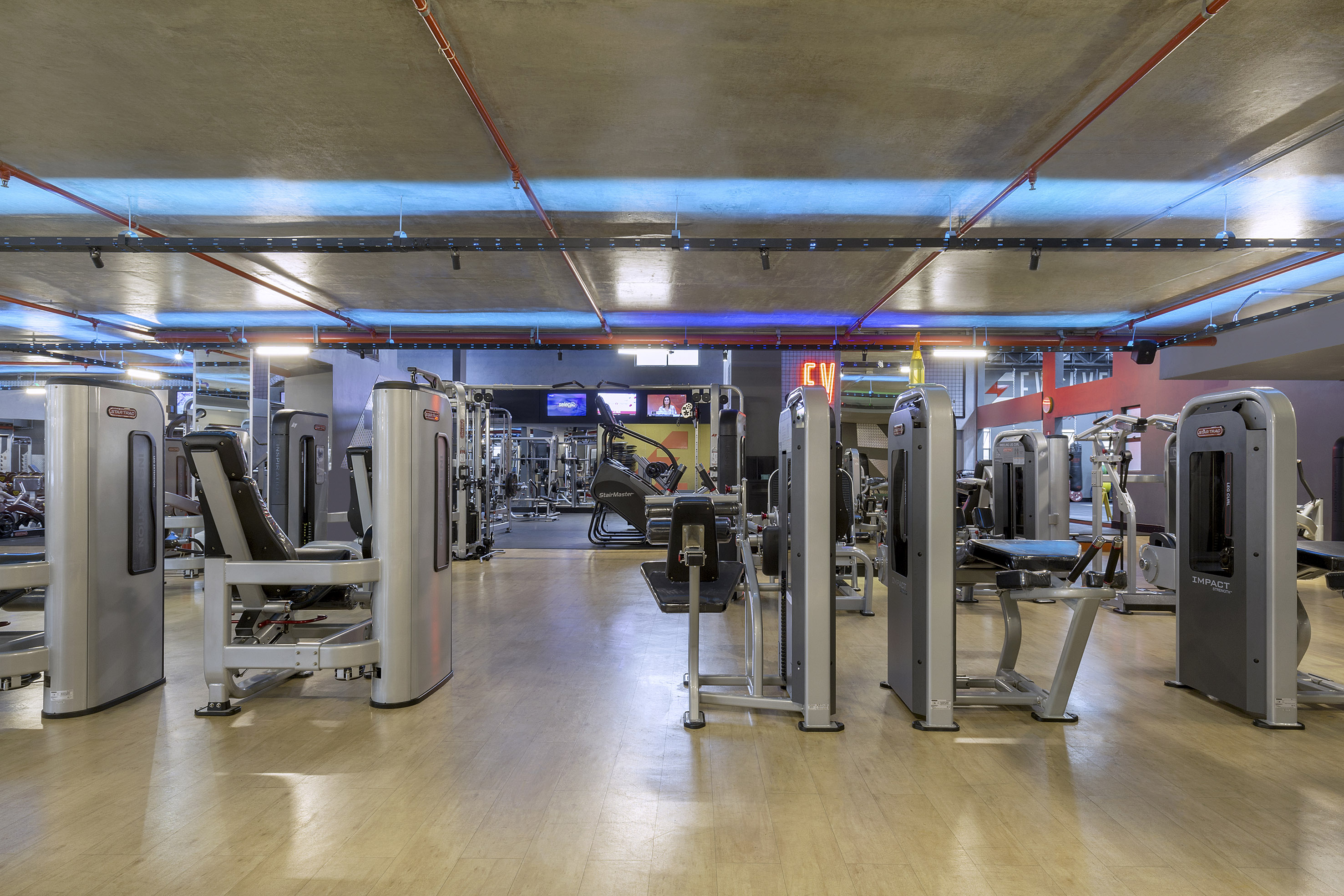 Gym interior with diverse strength and cardio workout machines under blue lighting.