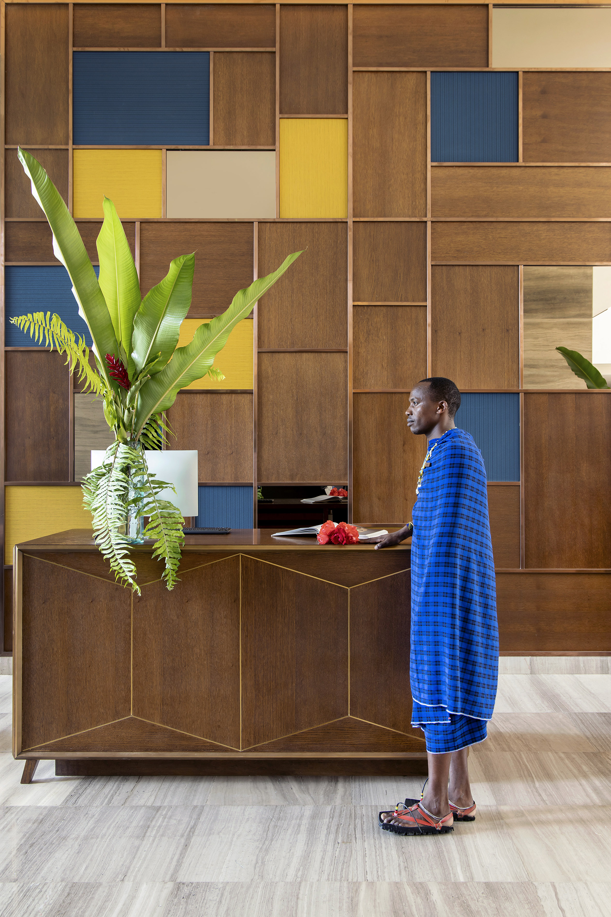 Maasai man at modern hotel reception desk with geometric wood wall and lush plant.