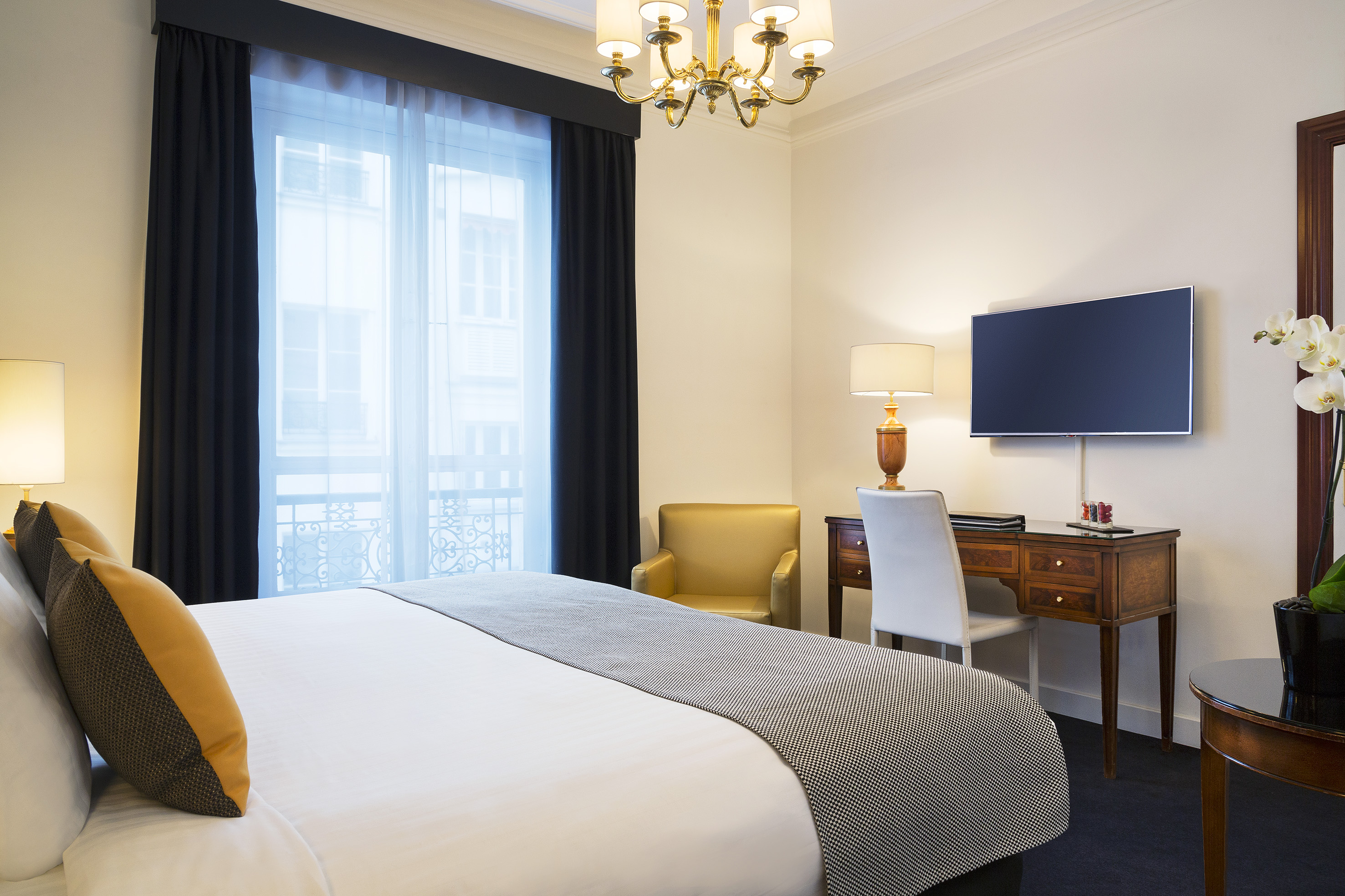 Hotel room with bed, desk, wall-mounted TV, and chandelier.