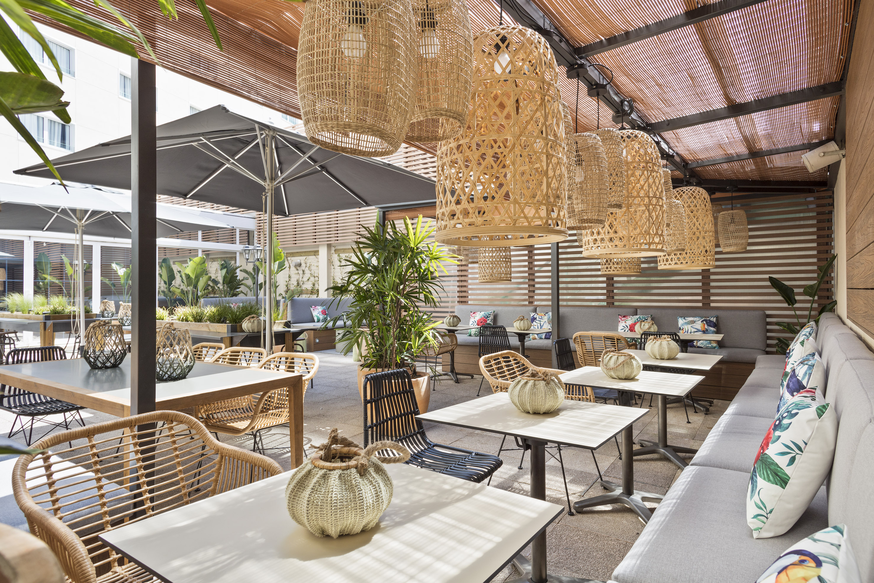 Outdoor restaurant patio with wicker chairs, rattan lamps, and tropical plants.