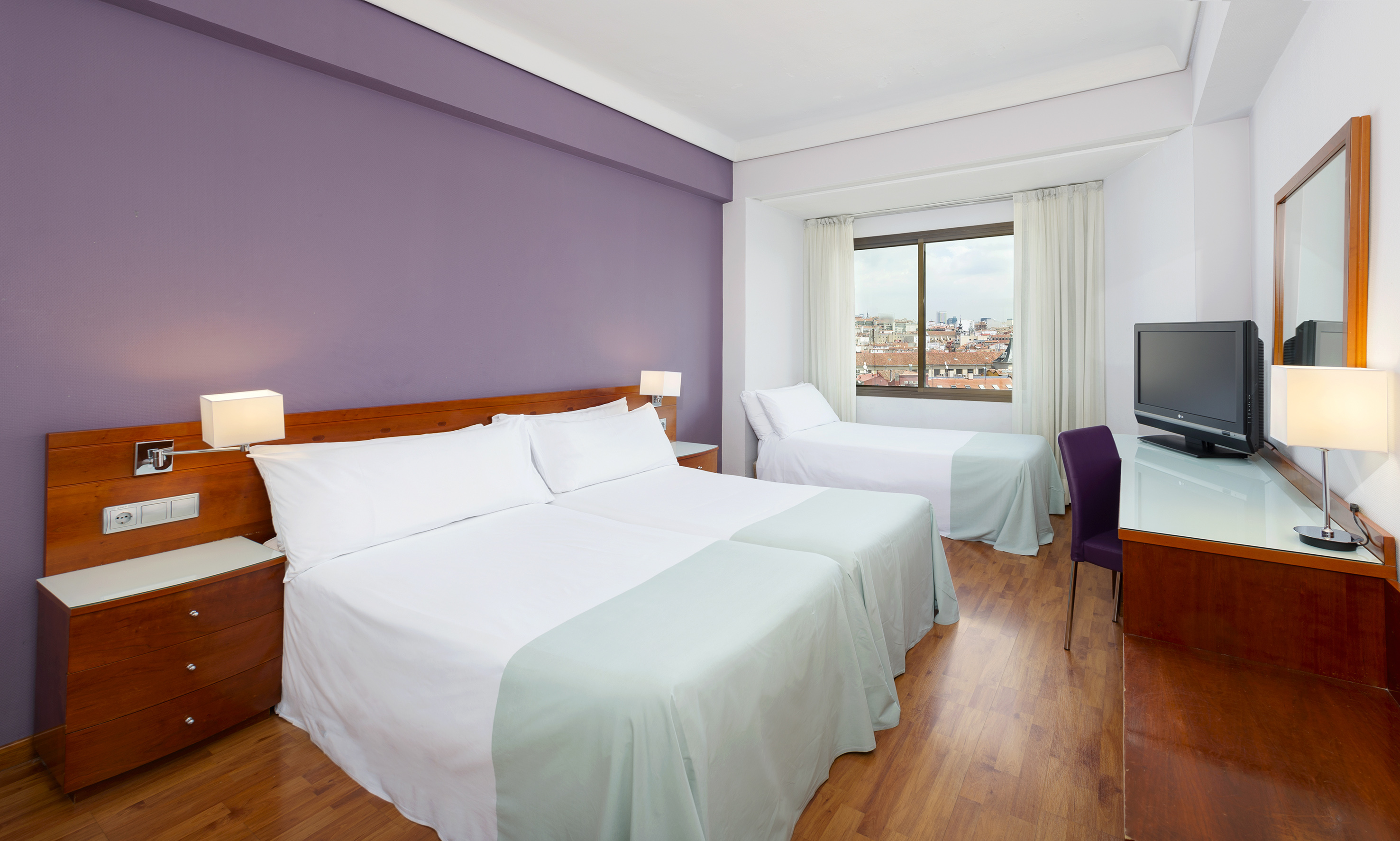 Hotel room with three beds, purple wall, city view, desk, and TV.