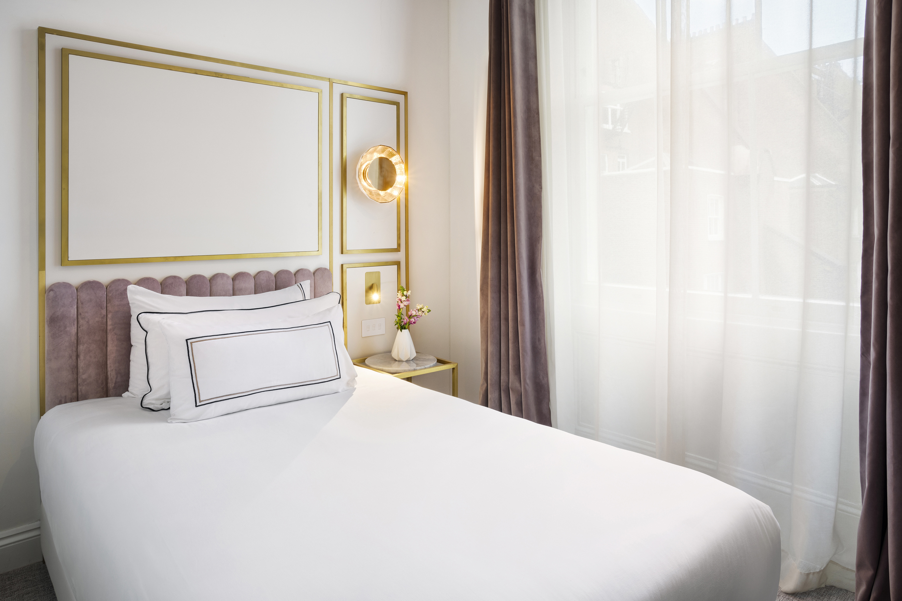 Chic hotel room bed with white linens, pink velvet headboard, gold decor.