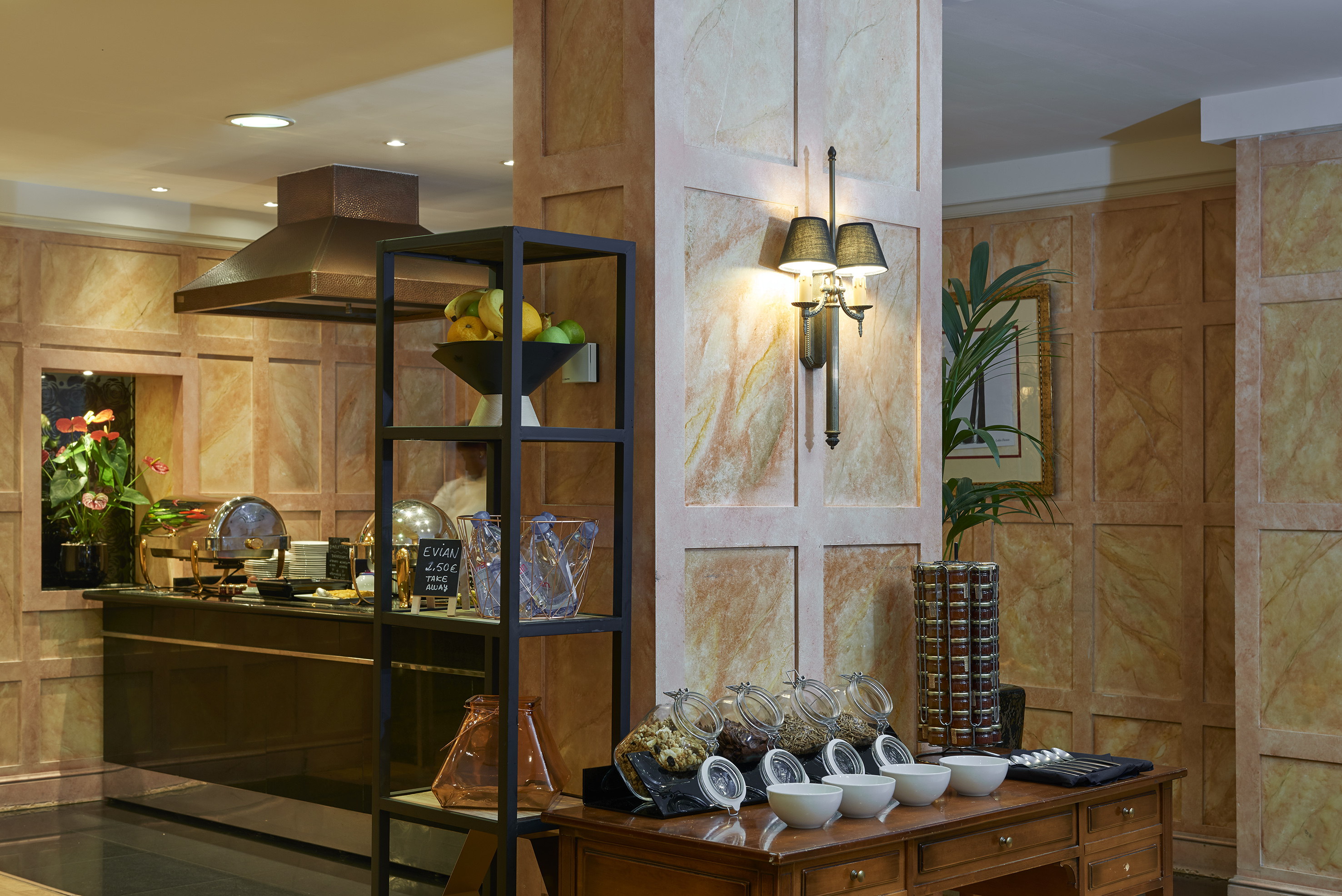 Hotel buffet breakfast station with cereals, fruits, hot food, and marbled walls.