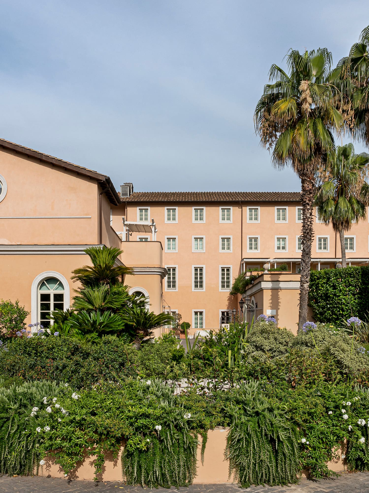 Grand Italian building exterior with palm trees and lush circular garden.