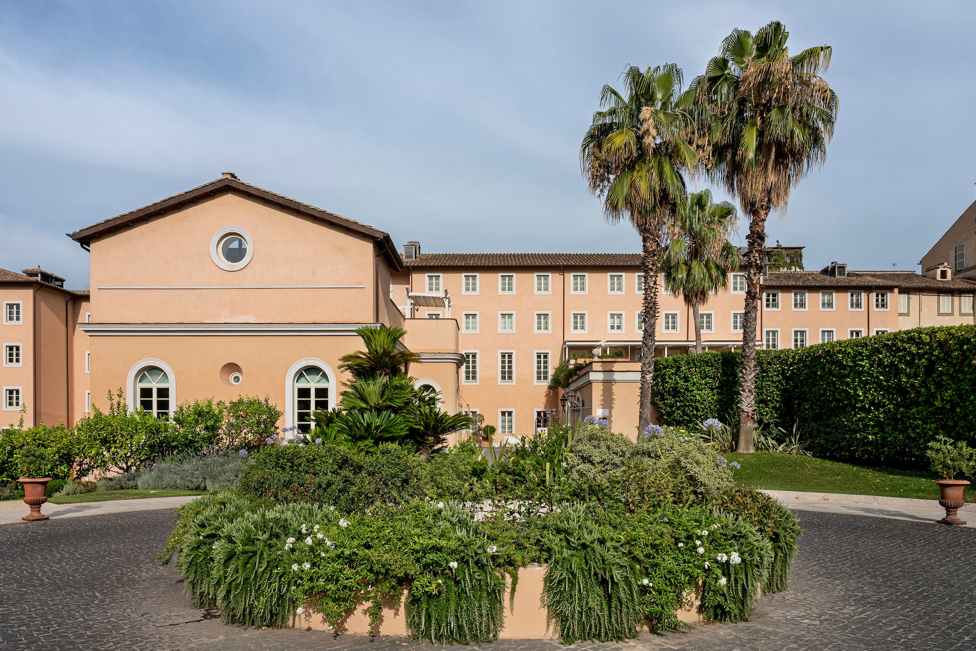 Peach Mediterranean building, lush garden, two tall palm trees, cobblestone path.