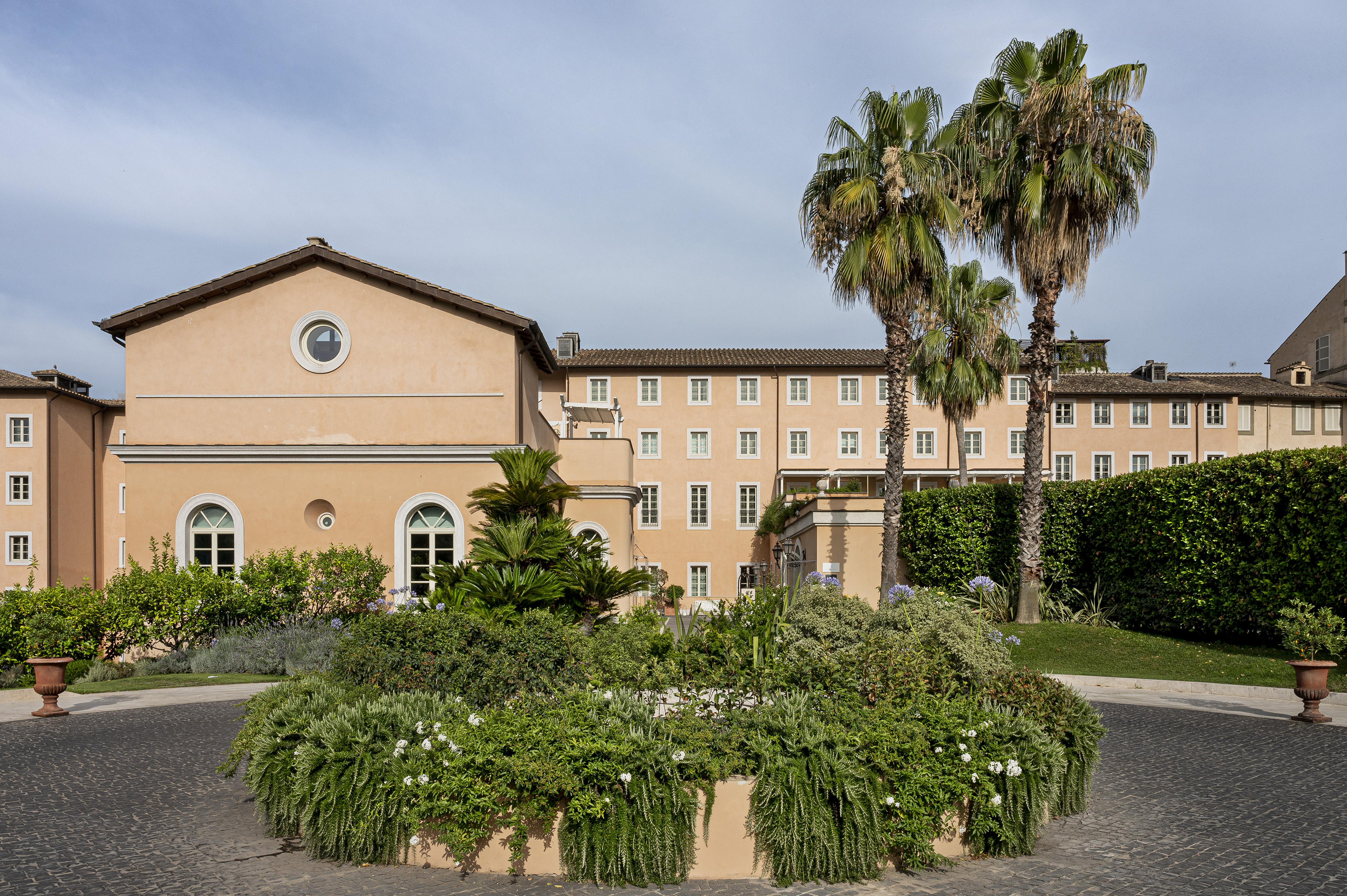 Peach Mediterranean building, lush garden, two tall palm trees, cobblestone path.