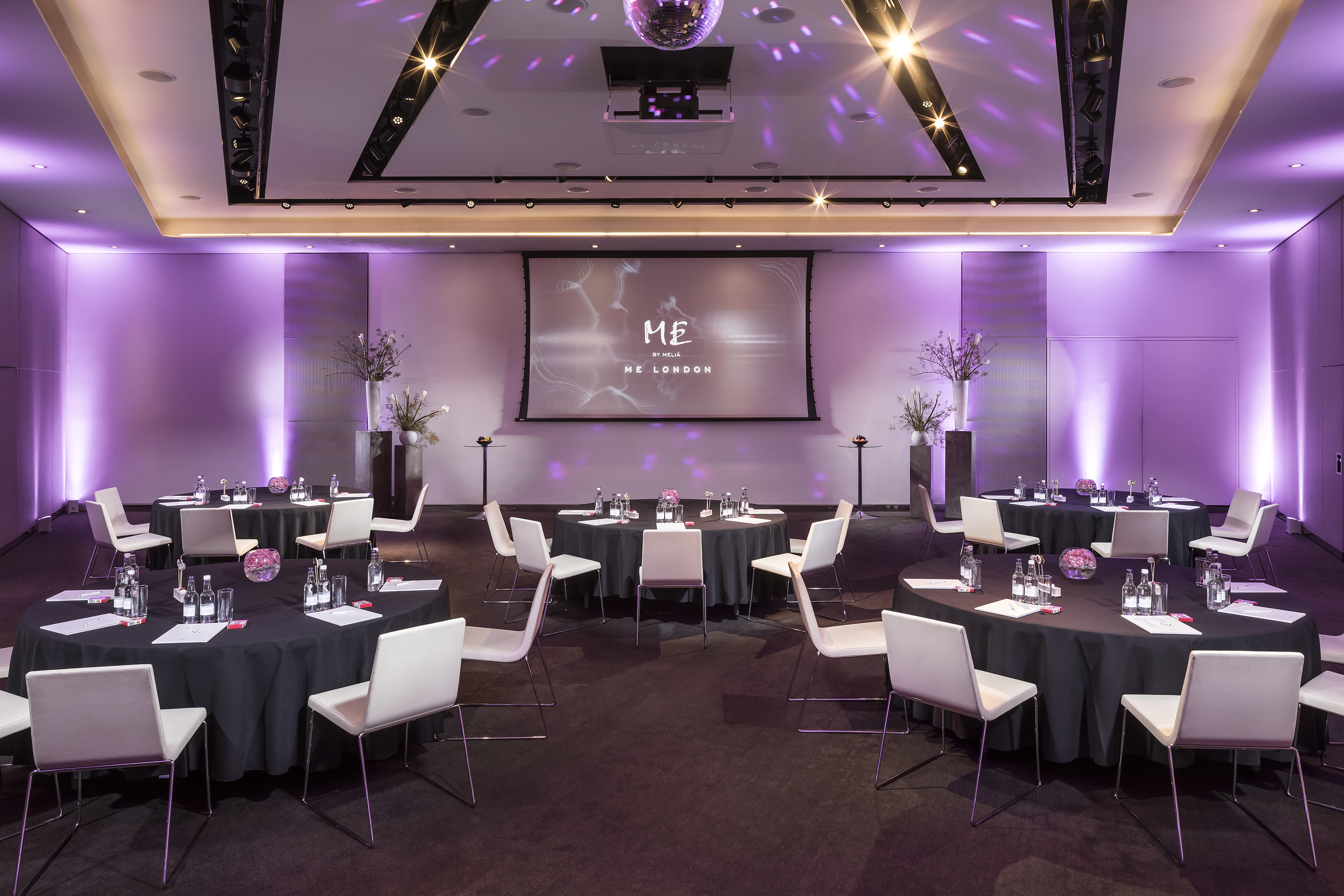 ME London event room with round tables, purple lighting, and projection screen.