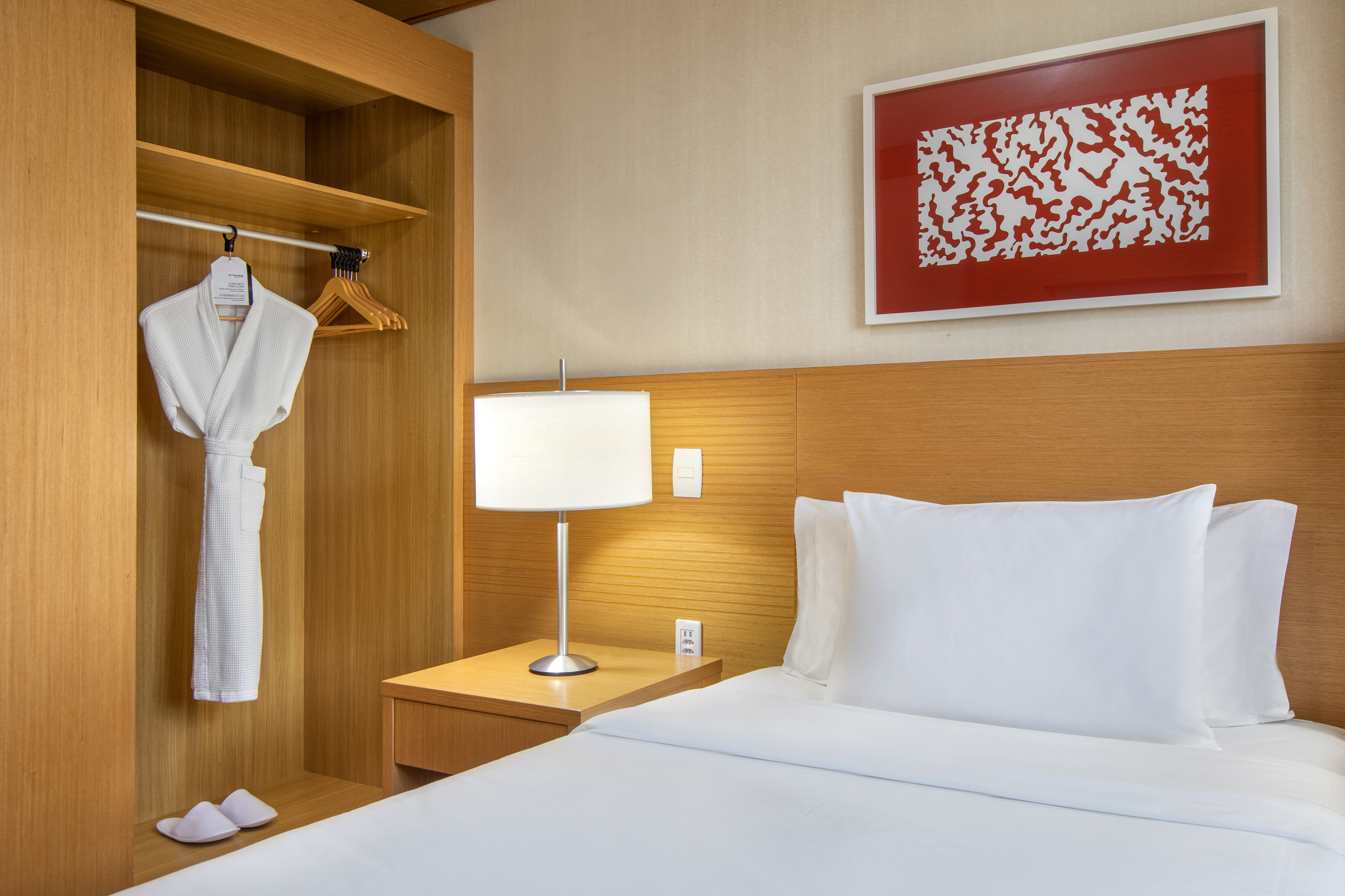 Modern hotel bedroom with white bed, wooden closet, bathrobe, and red art.