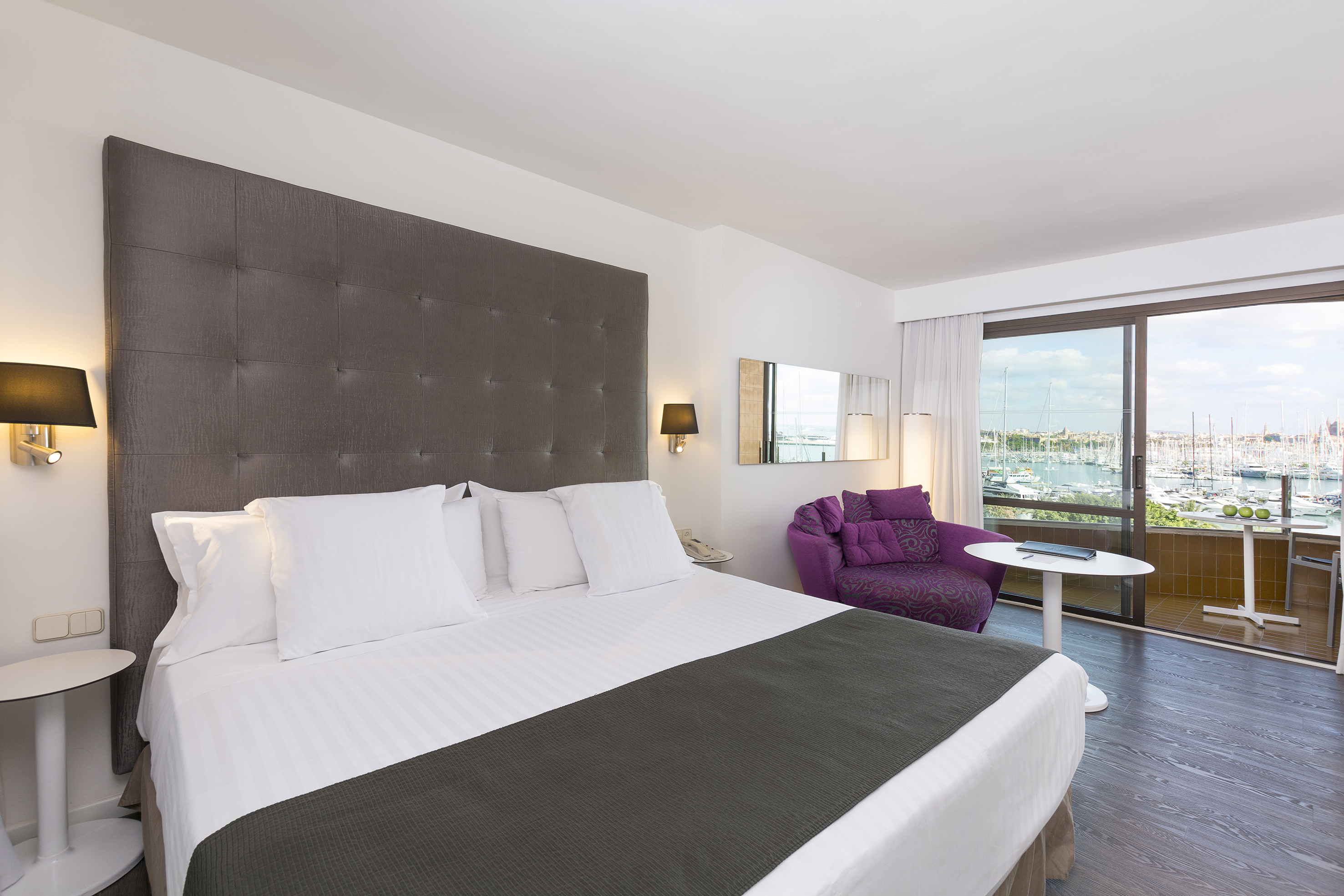Modern hotel bedroom with king bed, balcony marina view, and purple armchair.