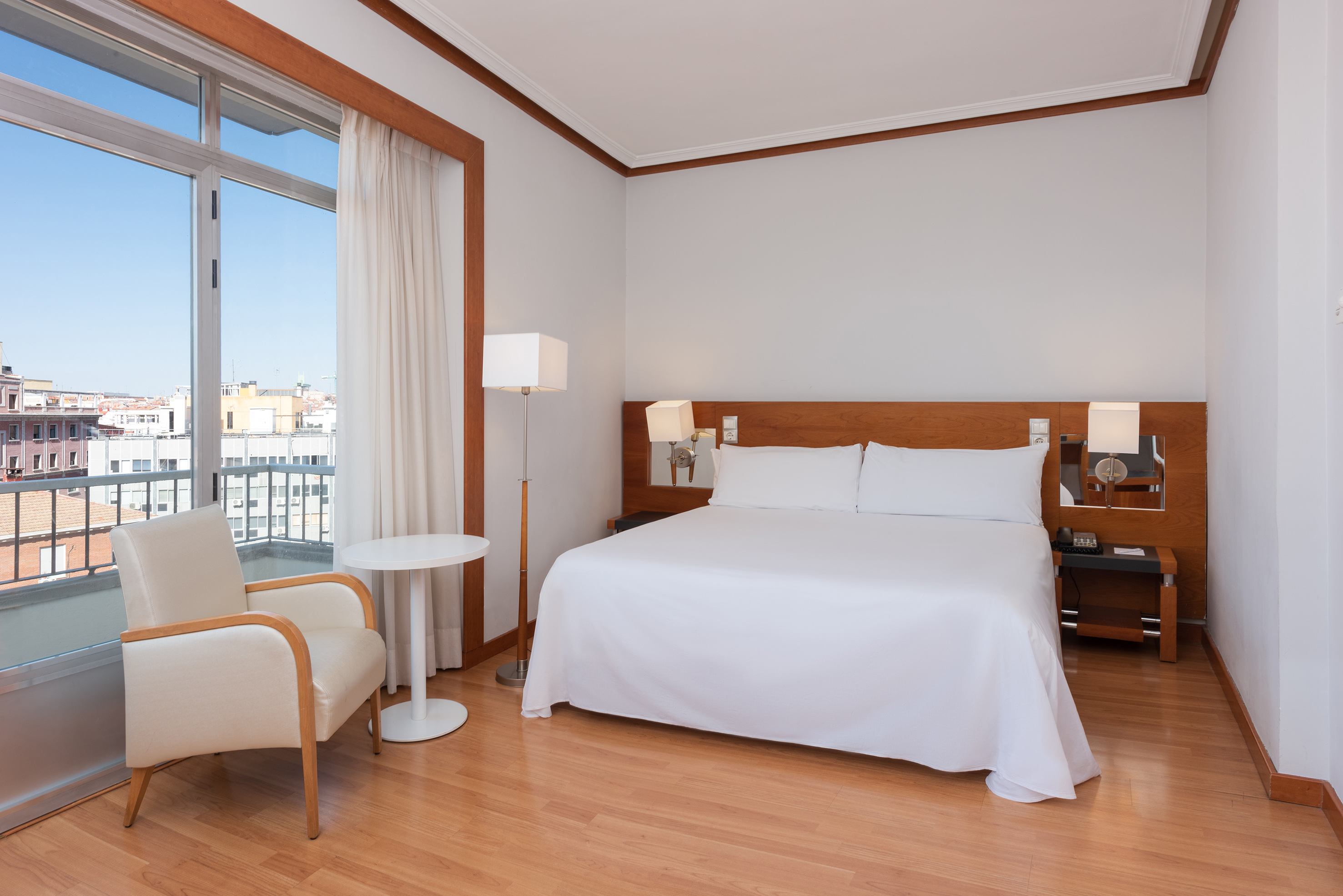 Modern guest room with white bed, armchair, city view window, and hardwood floor.